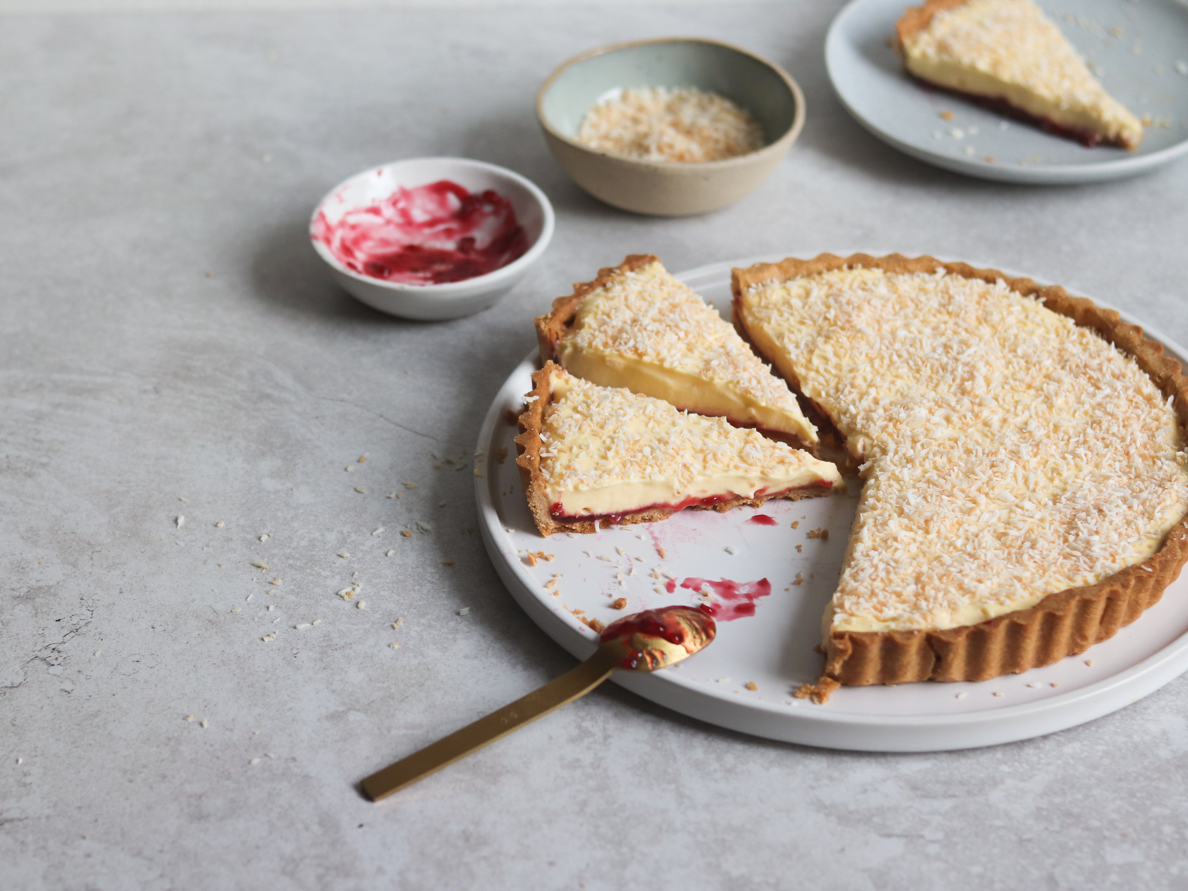 A manchester tart, with a slice cut out of it with a spoon covered in jam. Next to a bowl of jam and toasted coconut
