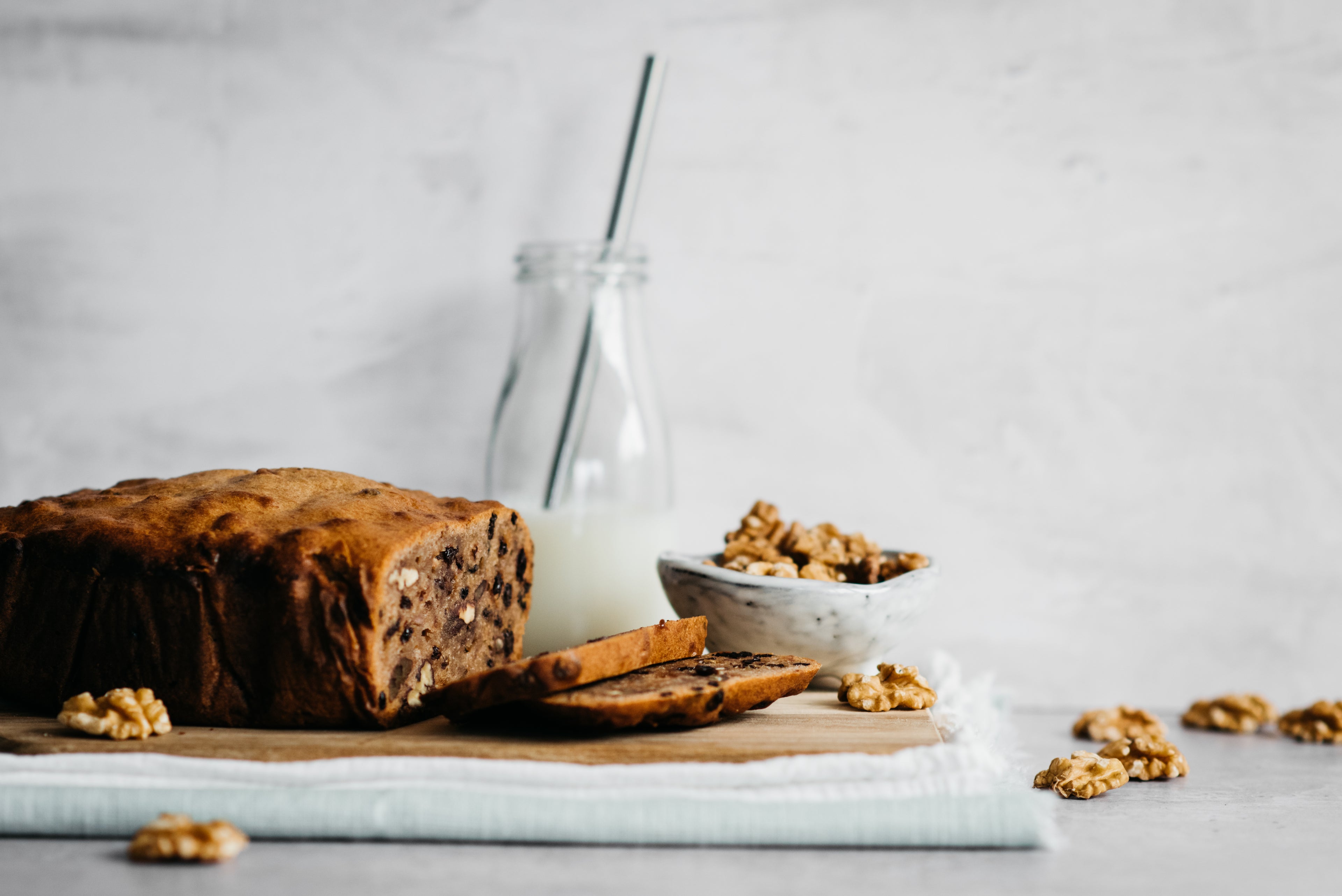 Loaf of banana bread with slices infront, milk jar in background and bowl of walnuts