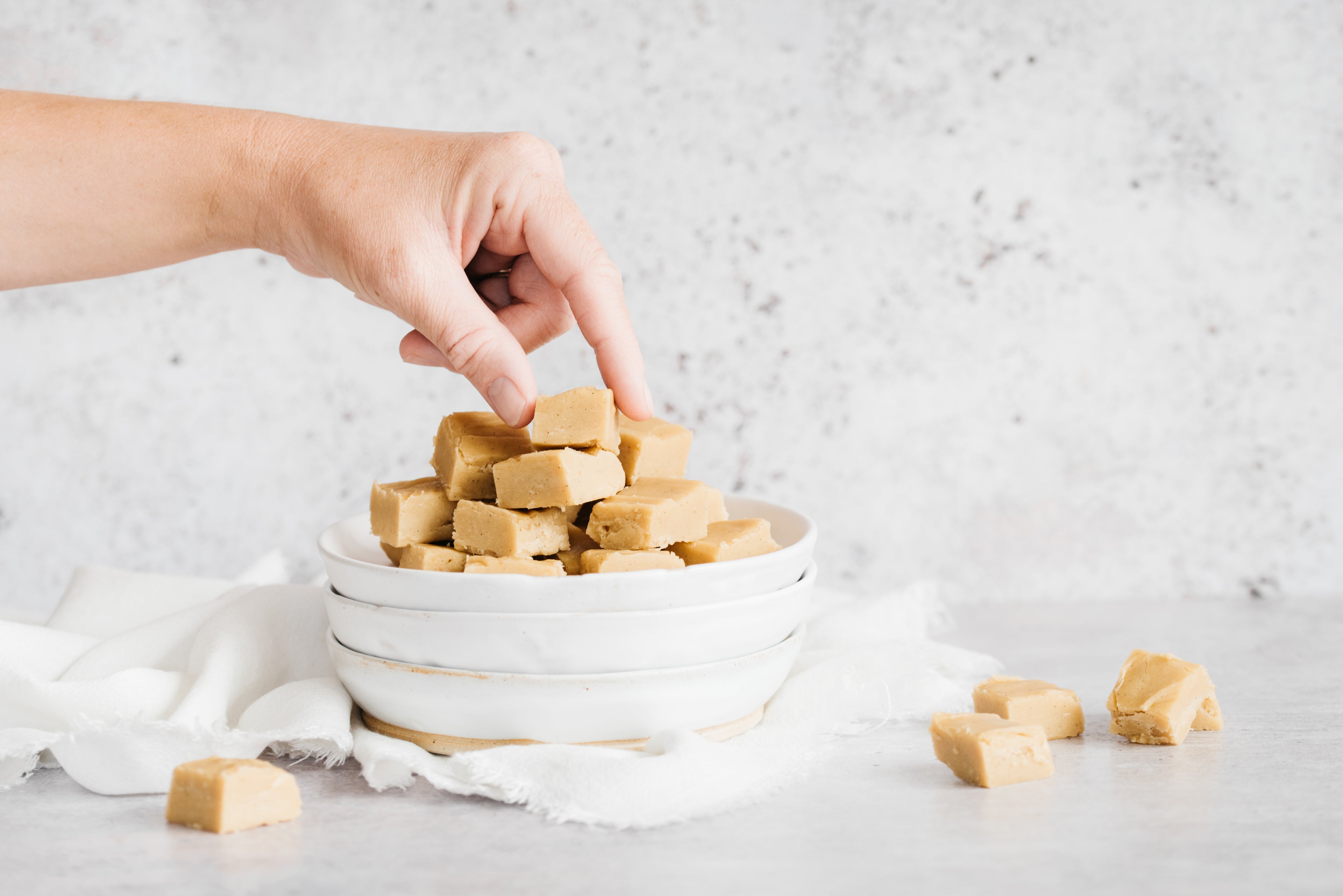 White bowl full of cubes of vanilla fudge. Hand reaching in to take one. 4 cubes of fudge in foreground