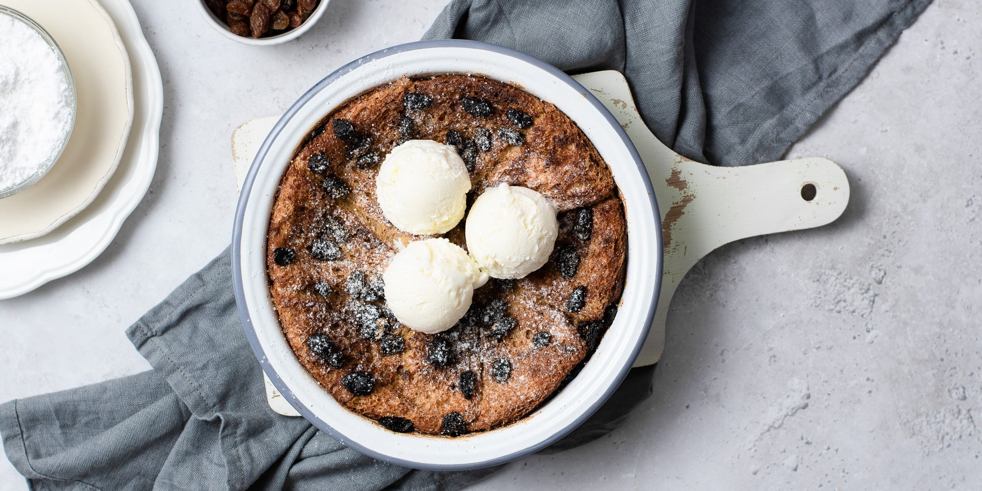 Classic Bread & Butter Pudding in white bowl with 3 scoops on ice cream on top. Bowl of icing sugar and raisins. On top of navy cloth