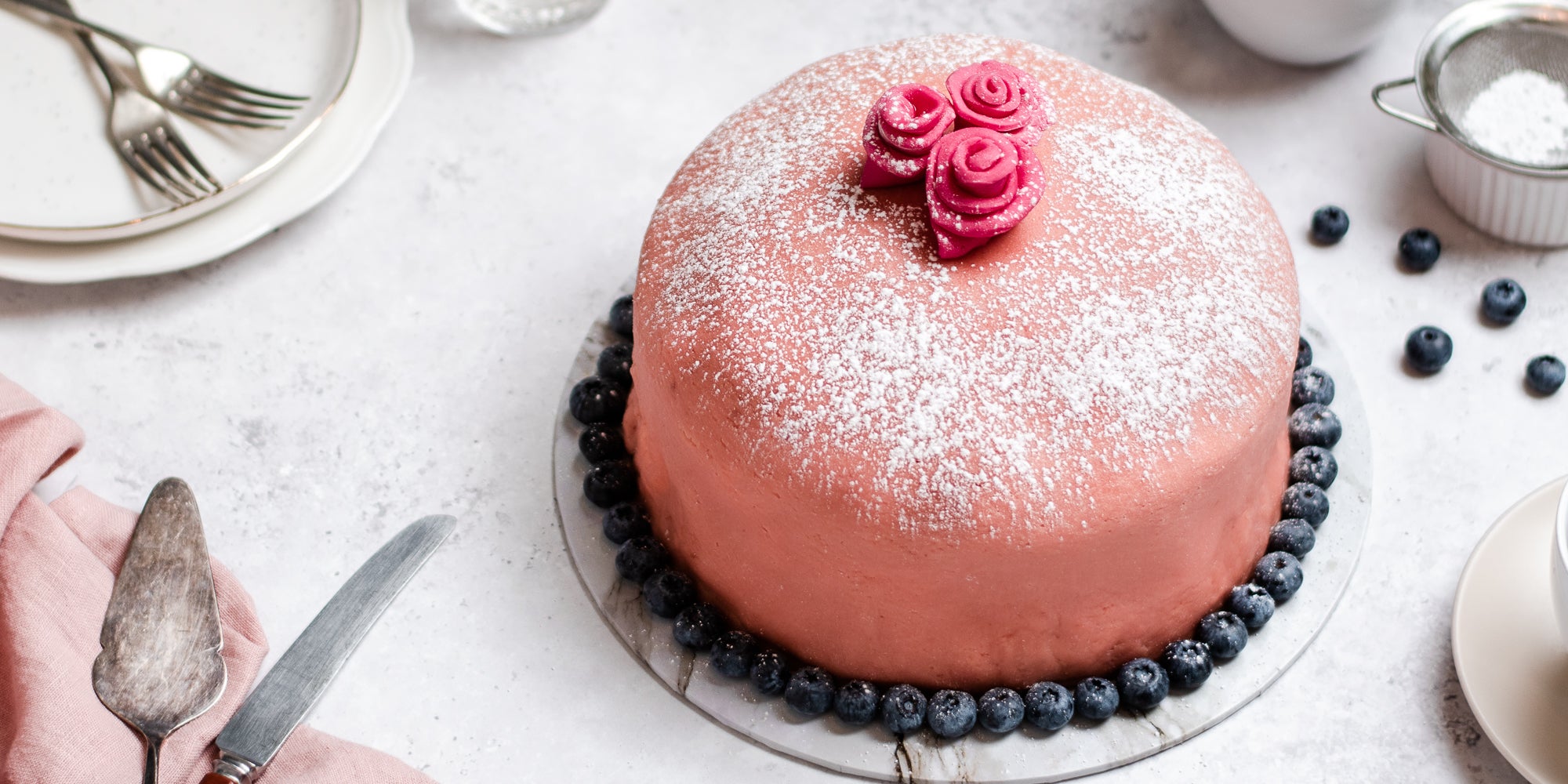 Close up view of Prinsesstarta surrounded with blueberries, and a cake knife to serve. Dusted with icing sugar and topped with fondant flowers