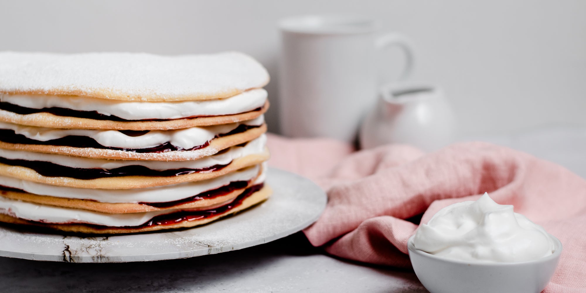 Close up of Danish Layer Cake on a marble serving plate, next to a bowl of whipped cream with a pink linen cloth in the background