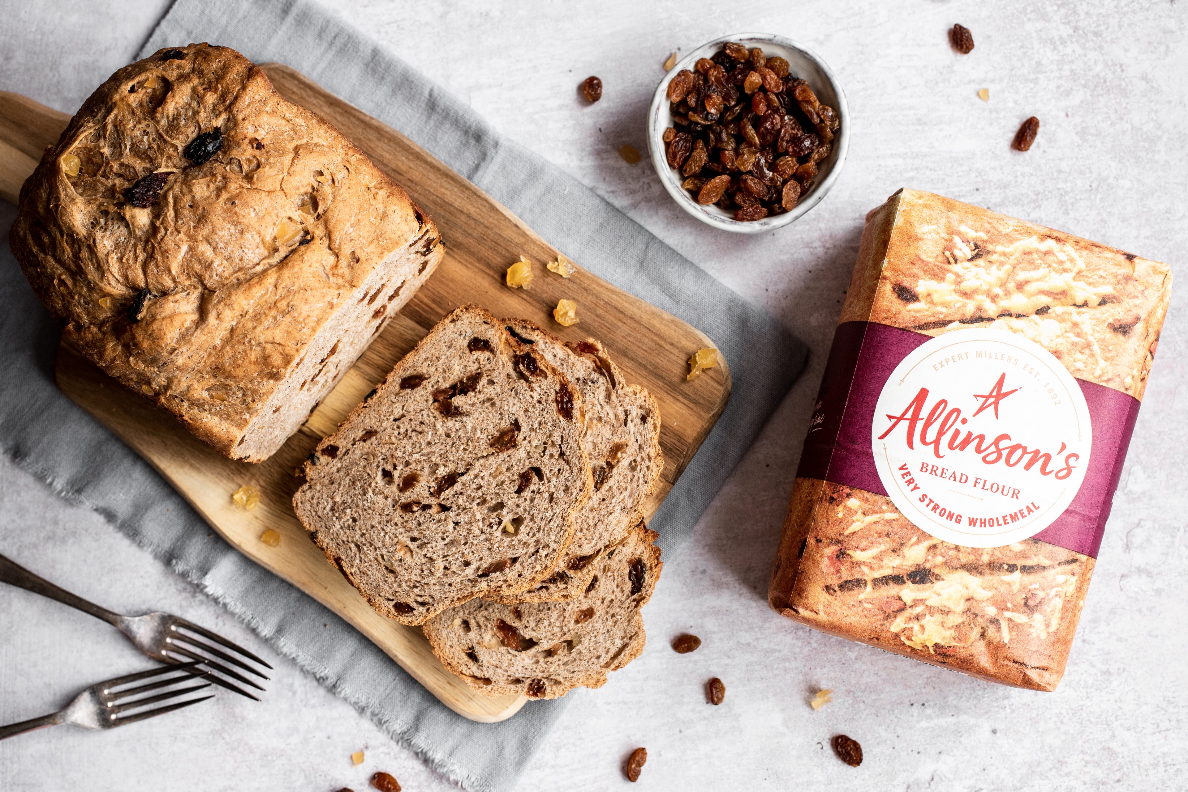 Overhead shot of bread with slices in front. Bowl of sultanas and flour pack. Two forks