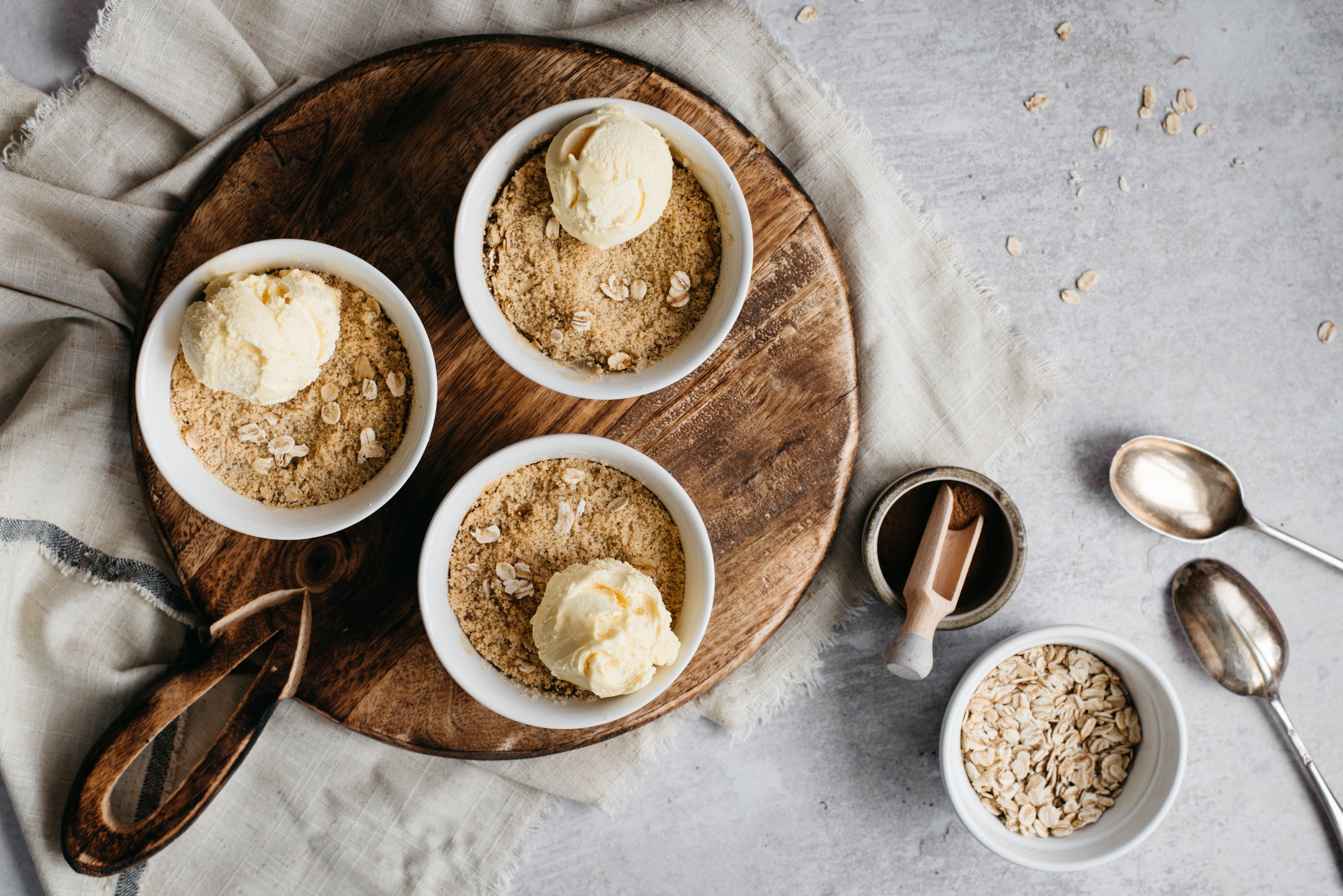 Mini Apple & Cinnamon Crumbles served in ramekin dishes, next to bowls of oats and served with a dollop of ice cream