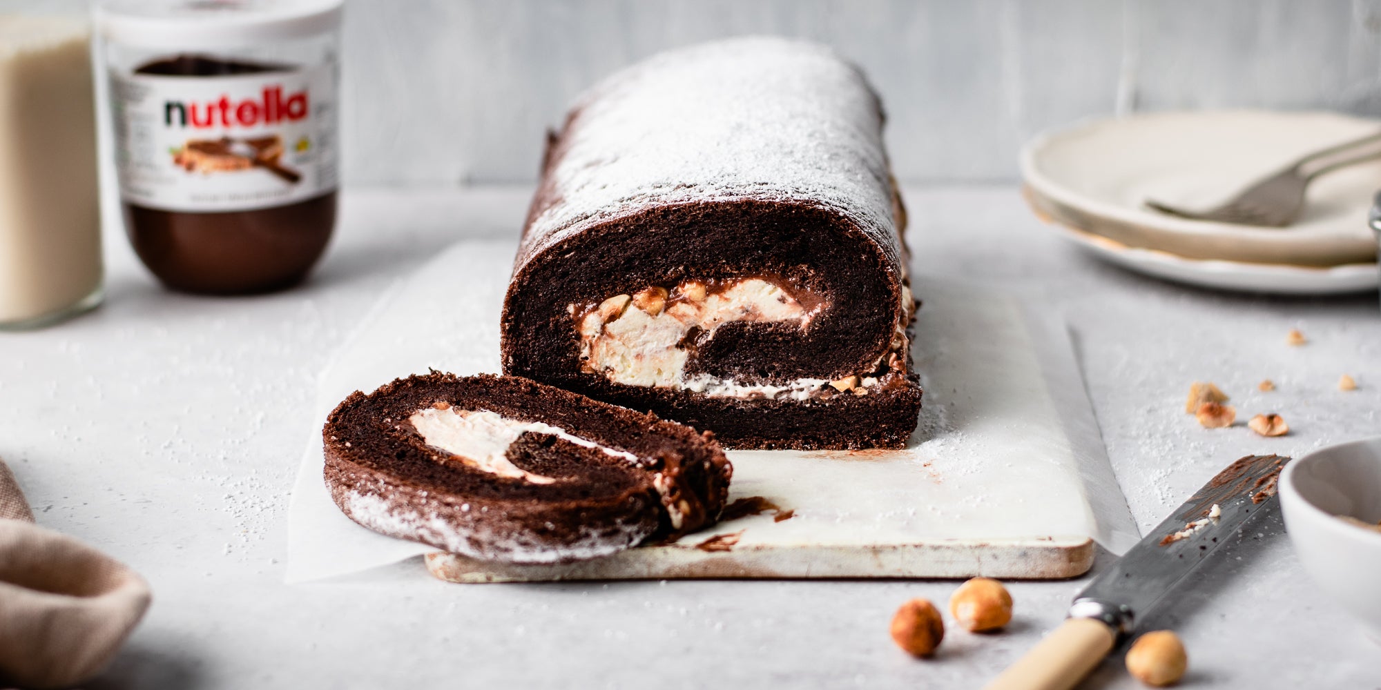 Chocolate Roulade sliced on a serving board, next to a knifes and scatters of hazelnuts