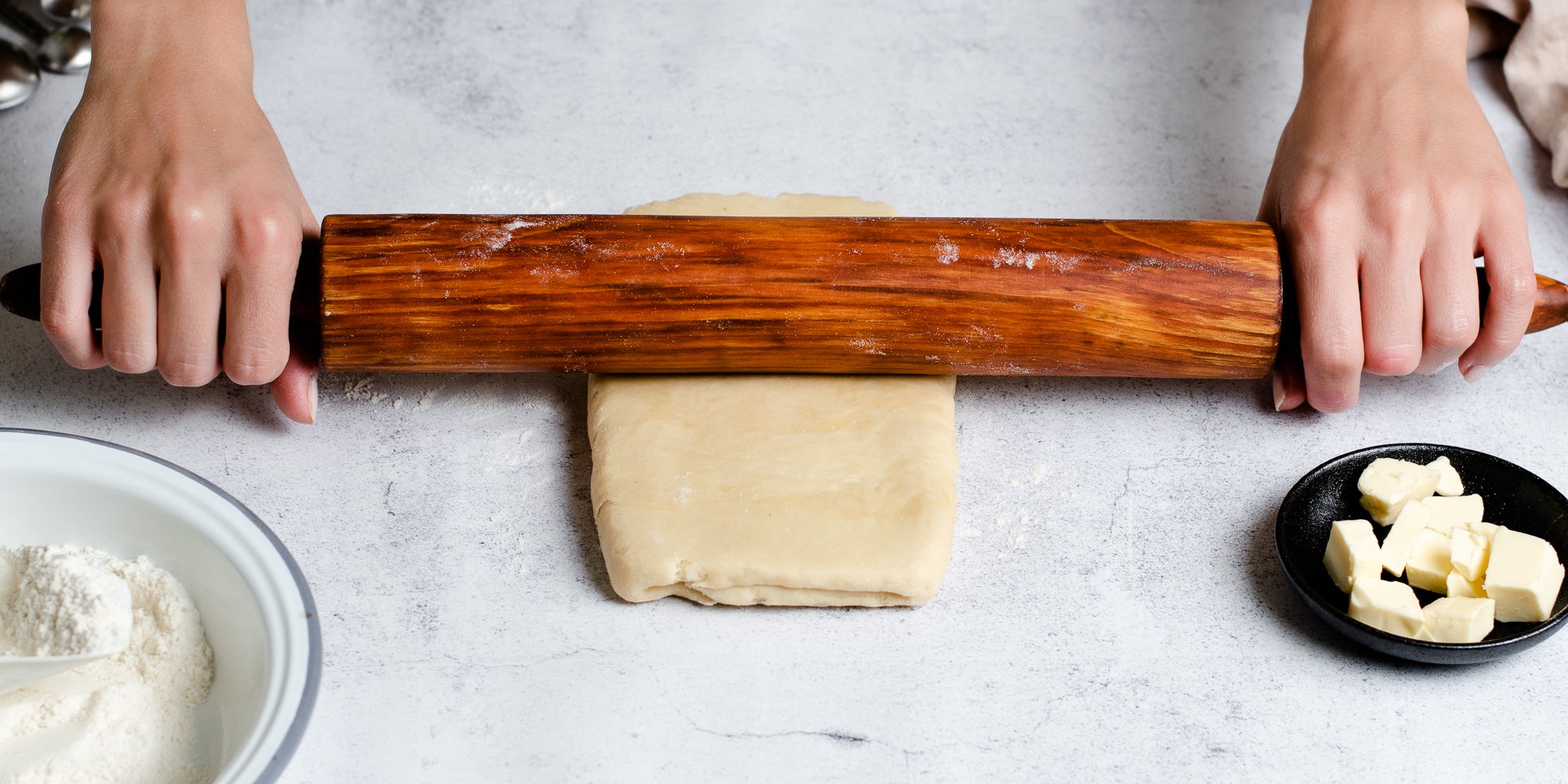 Hands rolling out Puff Pastry dough with a rolling pin, next to a dish of butter and a bowl of flour