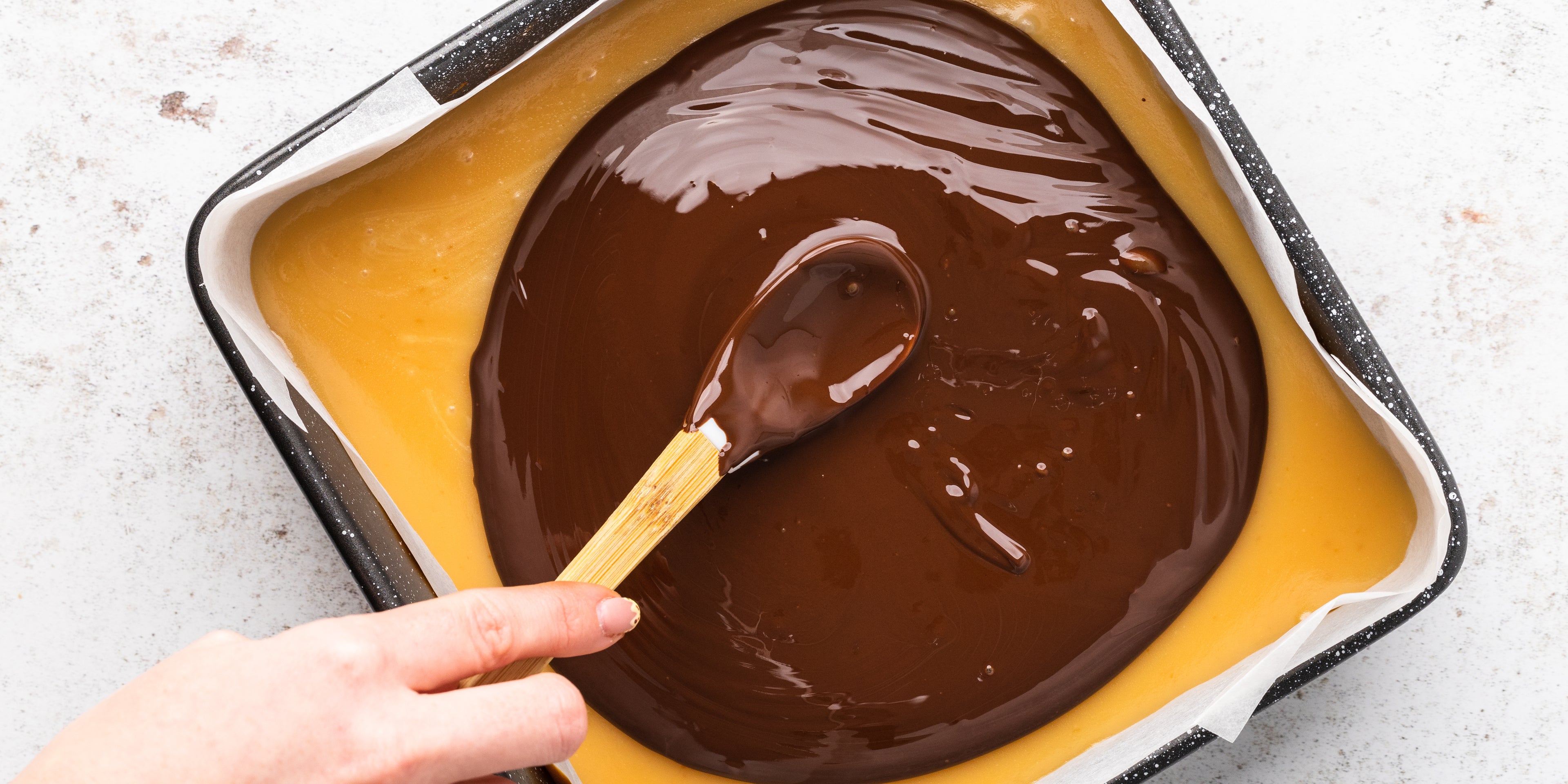Chocolate being spread over the top of caramel in a square baking tin