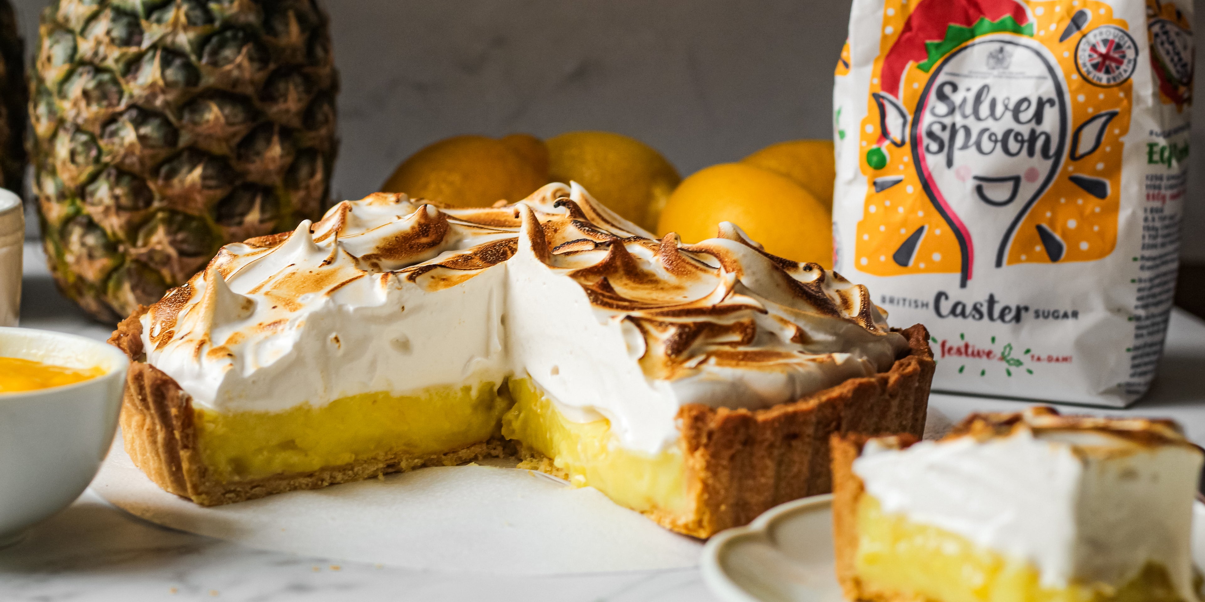 Vegan Pineapple Meringue Pie with a pineapple in the background, next to a bag of Silver Spoon Christmas edition caster sugar