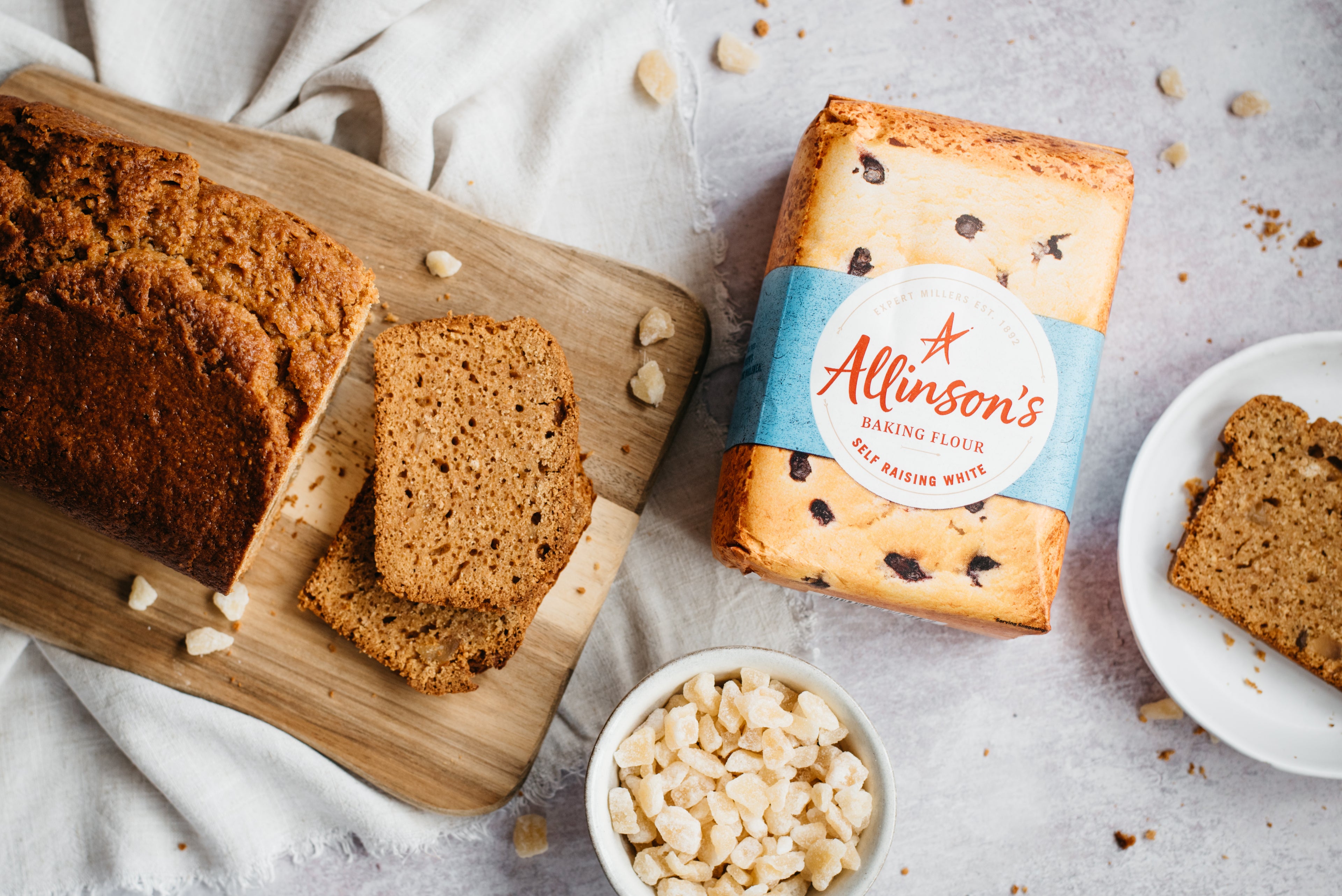 A flat lay of Gingerbread Loaf on a serving board, next to a bowl of ginger, bag of Allinson's flour and a slice of Gingerbread Loaf on a plate