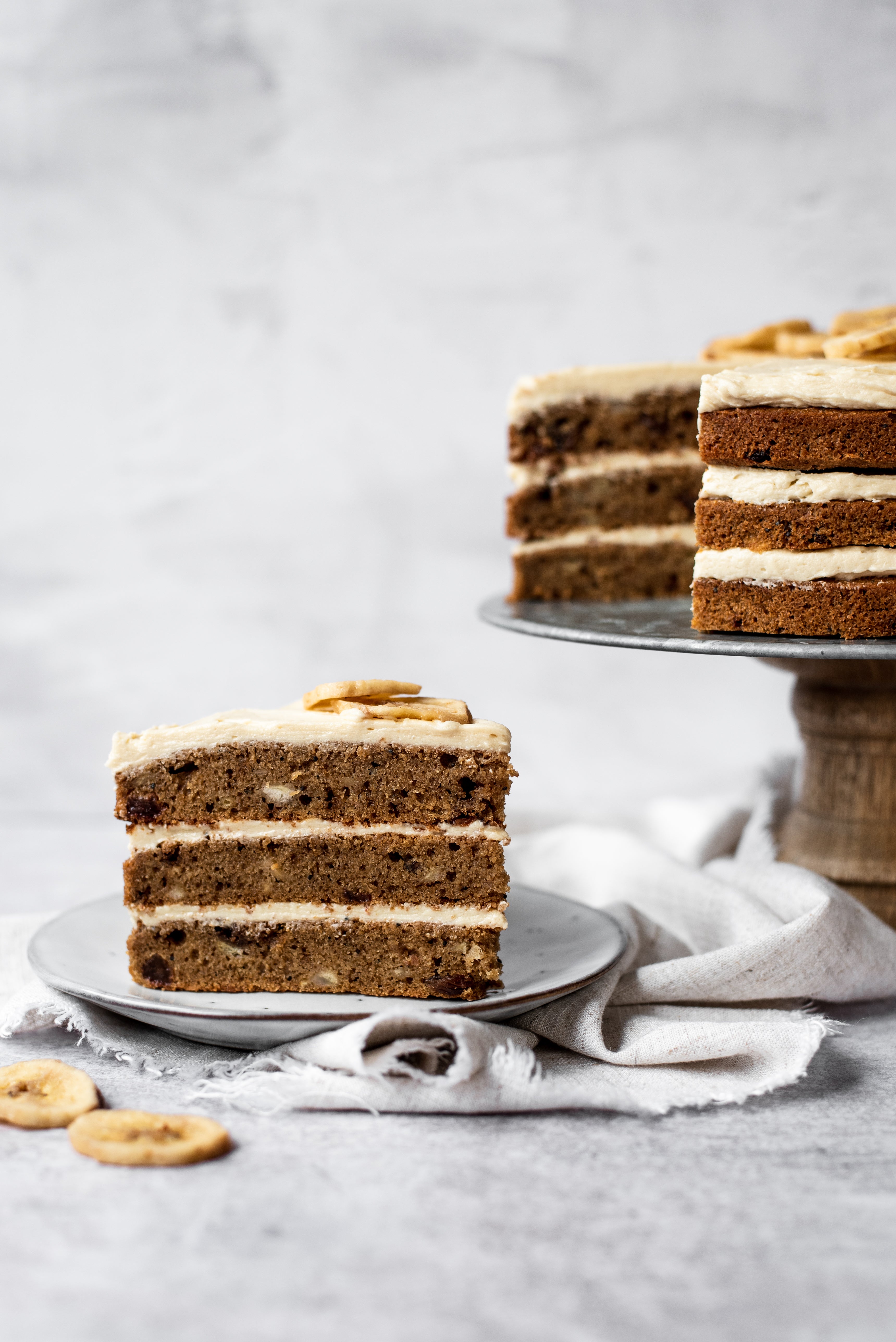 Slice of banana cake with 3 layers on a plate in front of remaining cake of cake stand