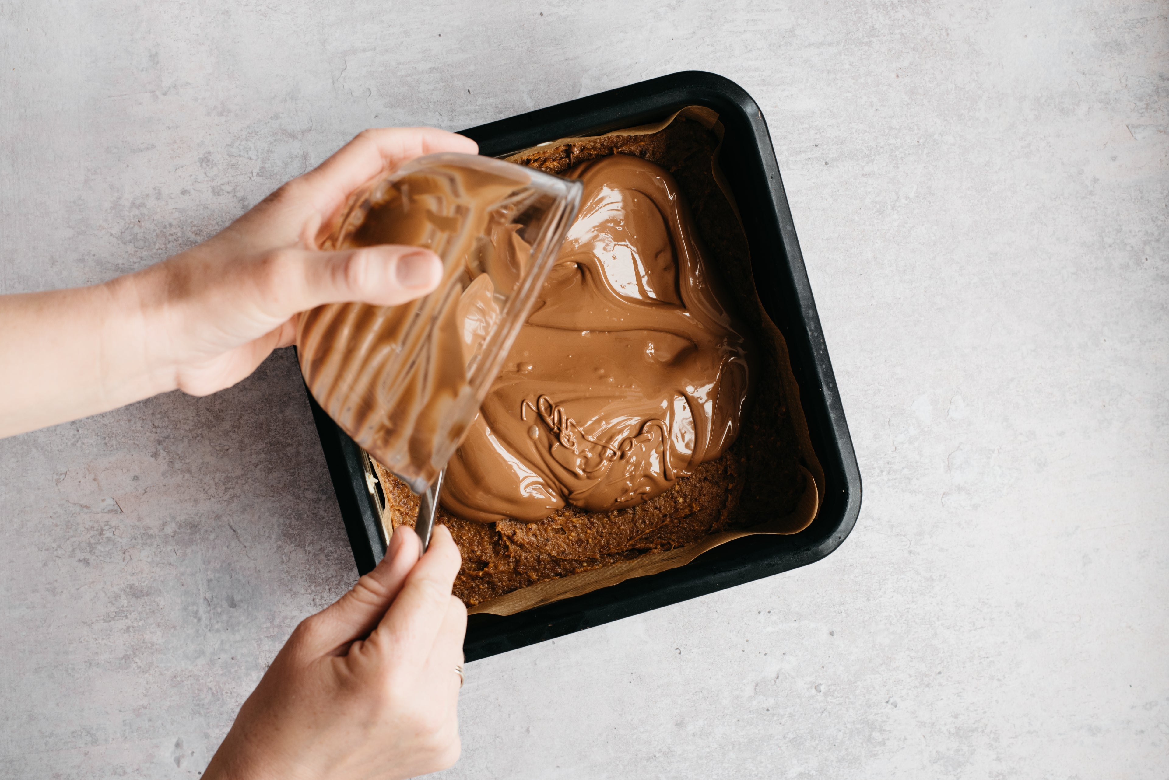 Hands holding a glass bowl spooning in the melted caramel into the baking tray to layer up the No-Bake Millionaire Bites