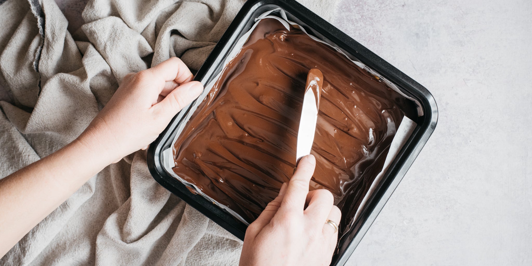 Melted chocolate being spread on top of a cake in a baking tin