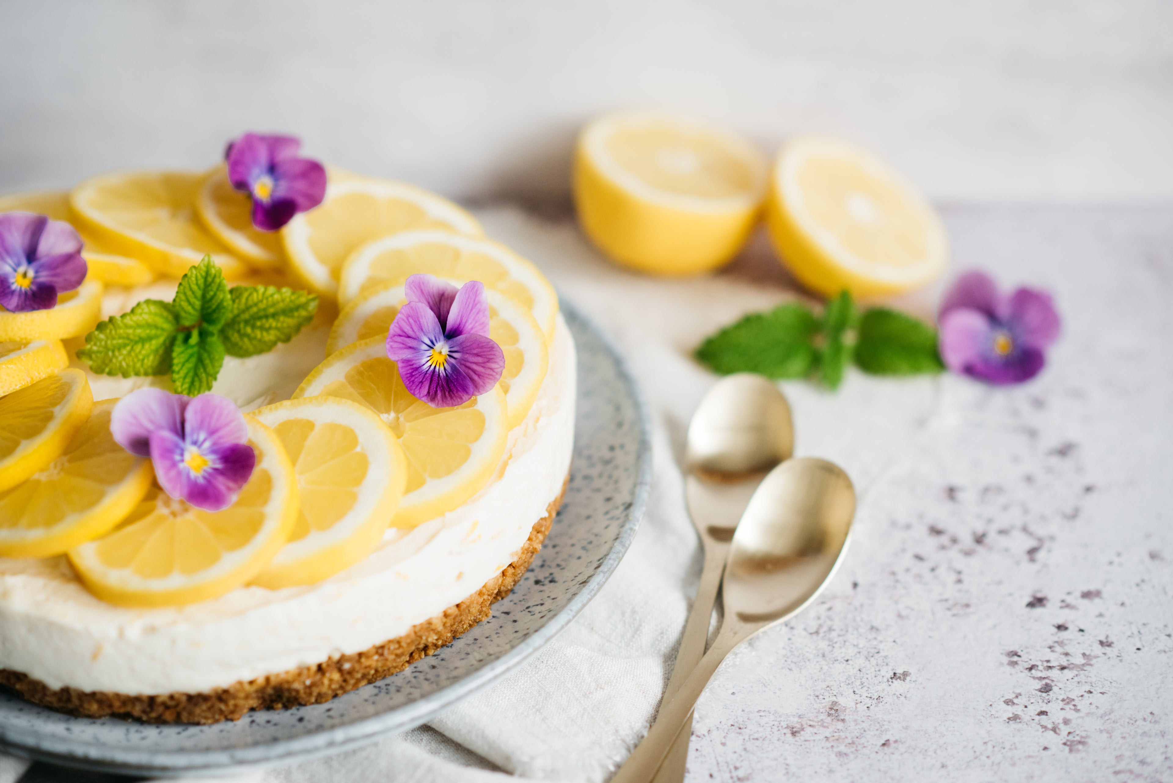 Close up of lemon cheesecake with purple flowers and lemon decoration and two spoons. Lemons in background