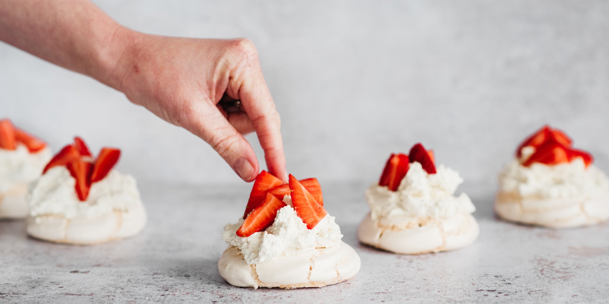 5 meringue nests topped with cream and strawberries in a line, with a hand reaching in adding a strawberry to the top of the centre meringue