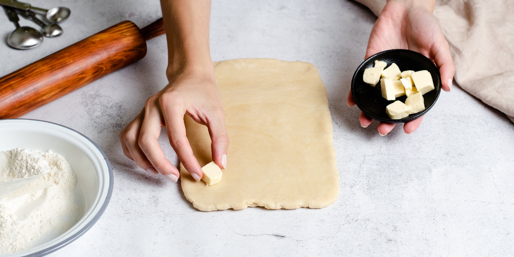 Hand laying out pieces of butter on rolled out Puff Pastry dough, ready to fold and roll out, next to a rolling pin and bowl of flour