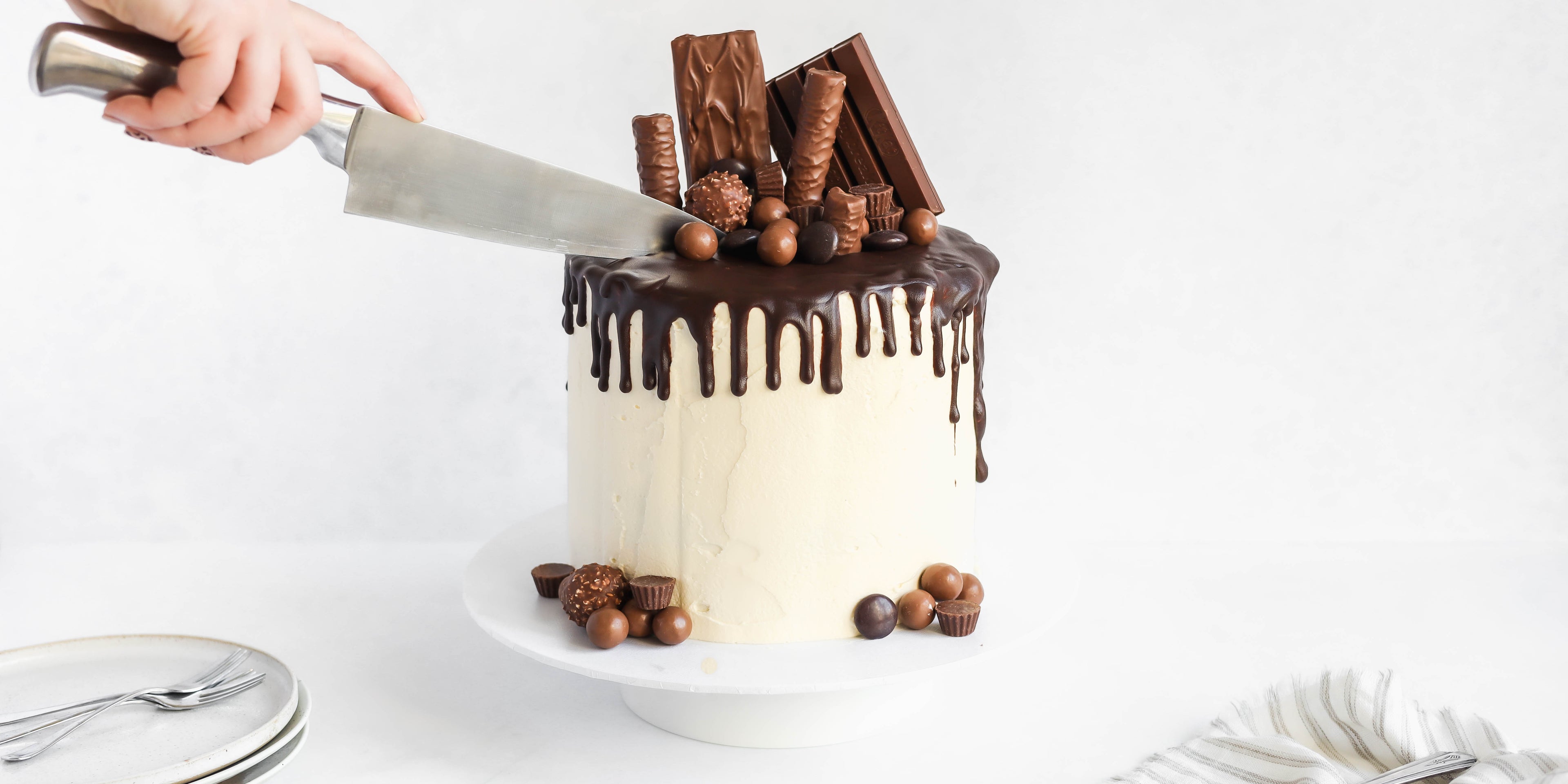 Ultimate Birthday Drip Cake with a hand reaching to slice into the cake with a knife. Topped with chocolate goodies