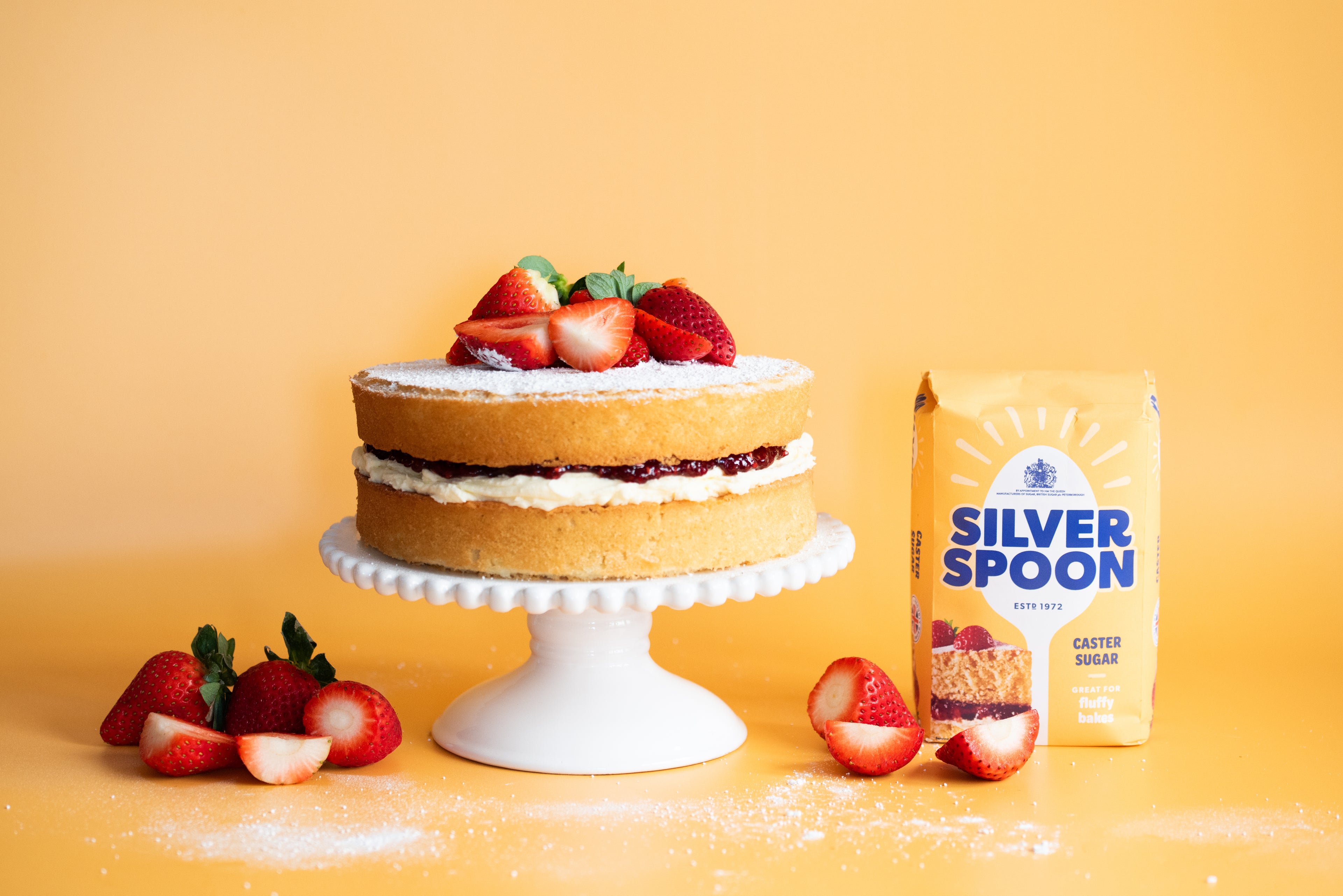 A victoria sponge cake topped with strawberries on a white cake stand beside strawberries and a pack of Silver Spoon sugar