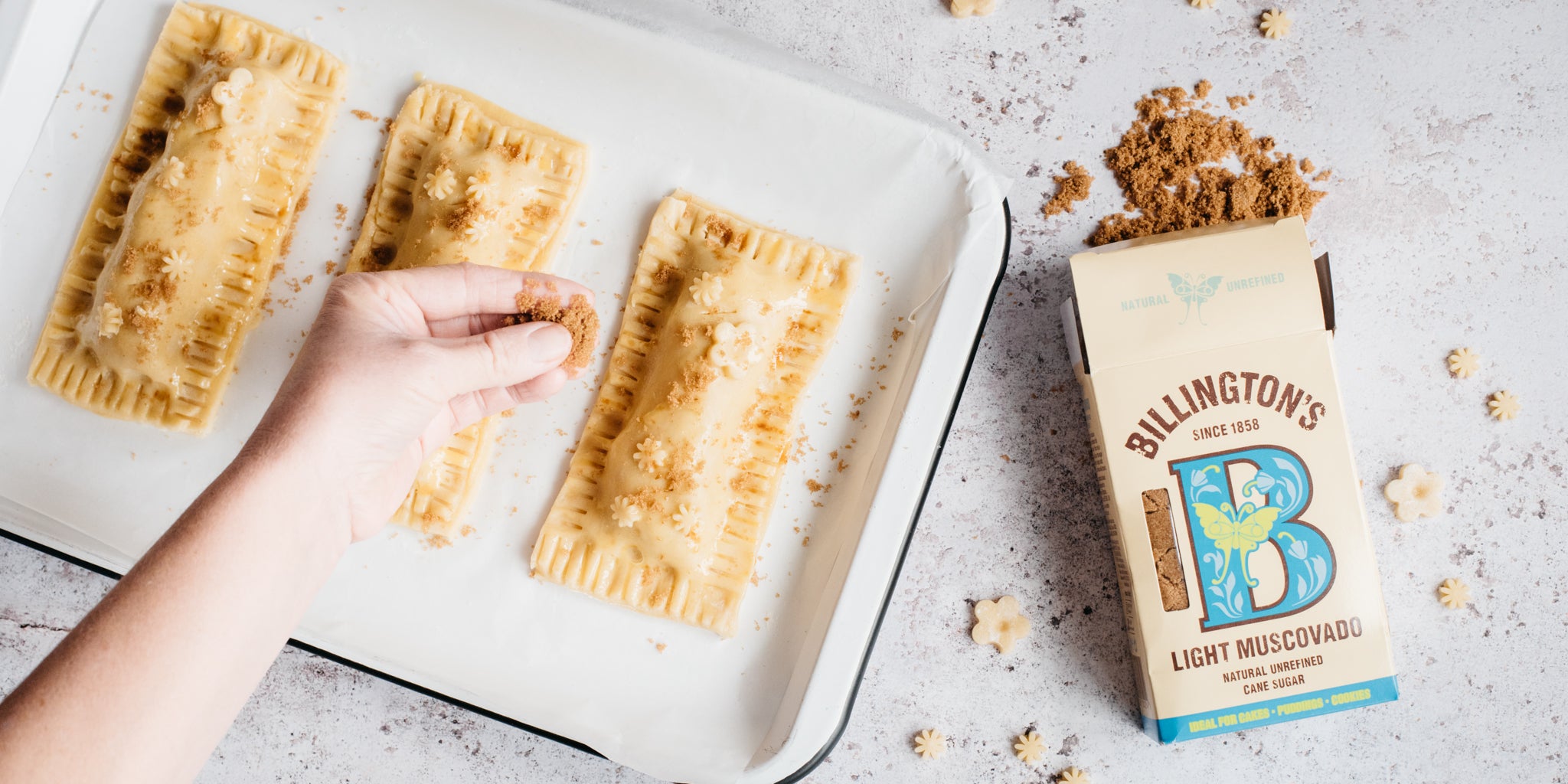 Apple pie pockets on a baking tray being sprinkled with sugar. Sugar pack open beside it