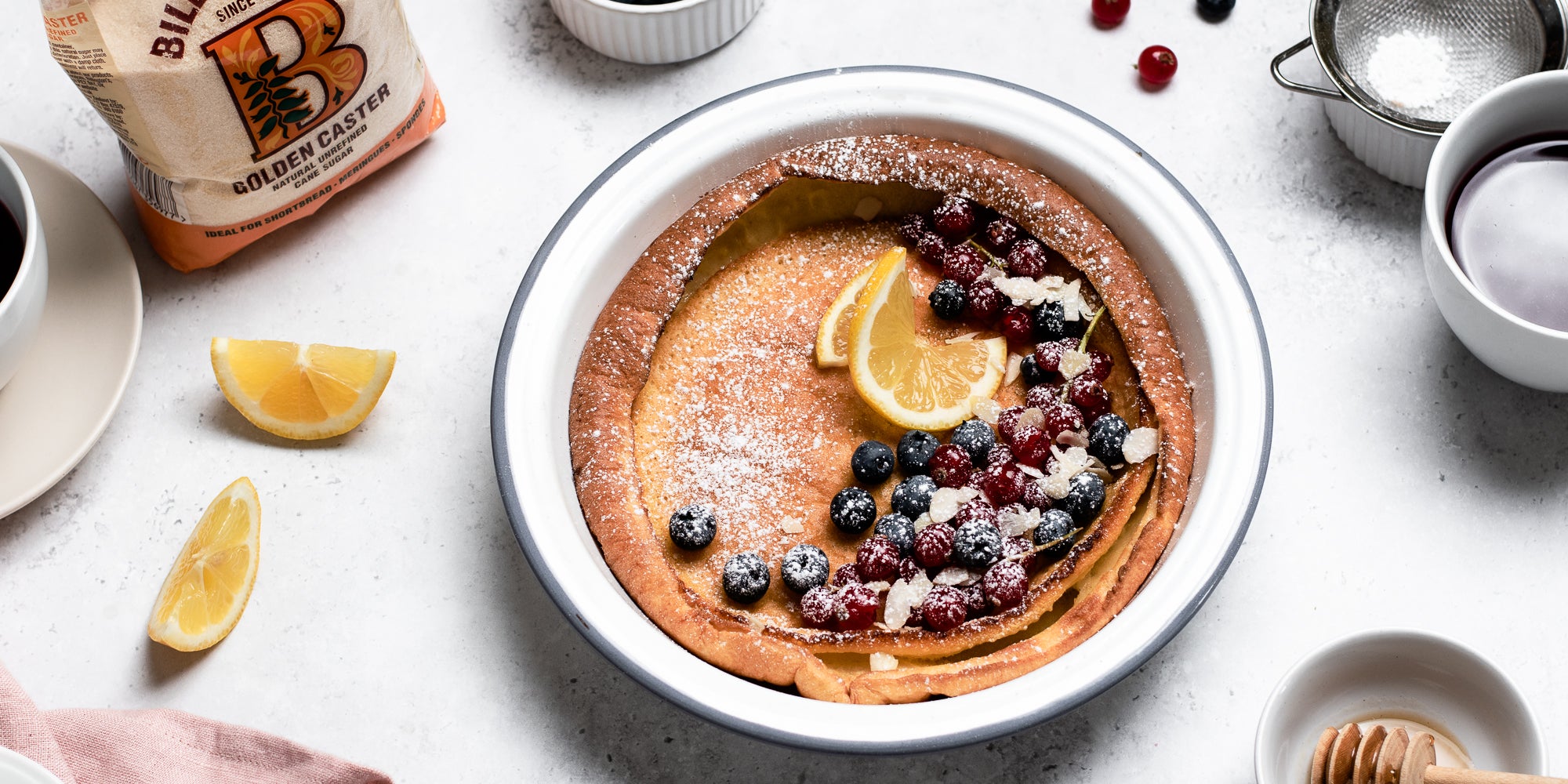 Close up of Dutch Baby Pancakes drizzled in honey, topped with berries and lemon next to a pack of Billington's Golden Caster Sugar 