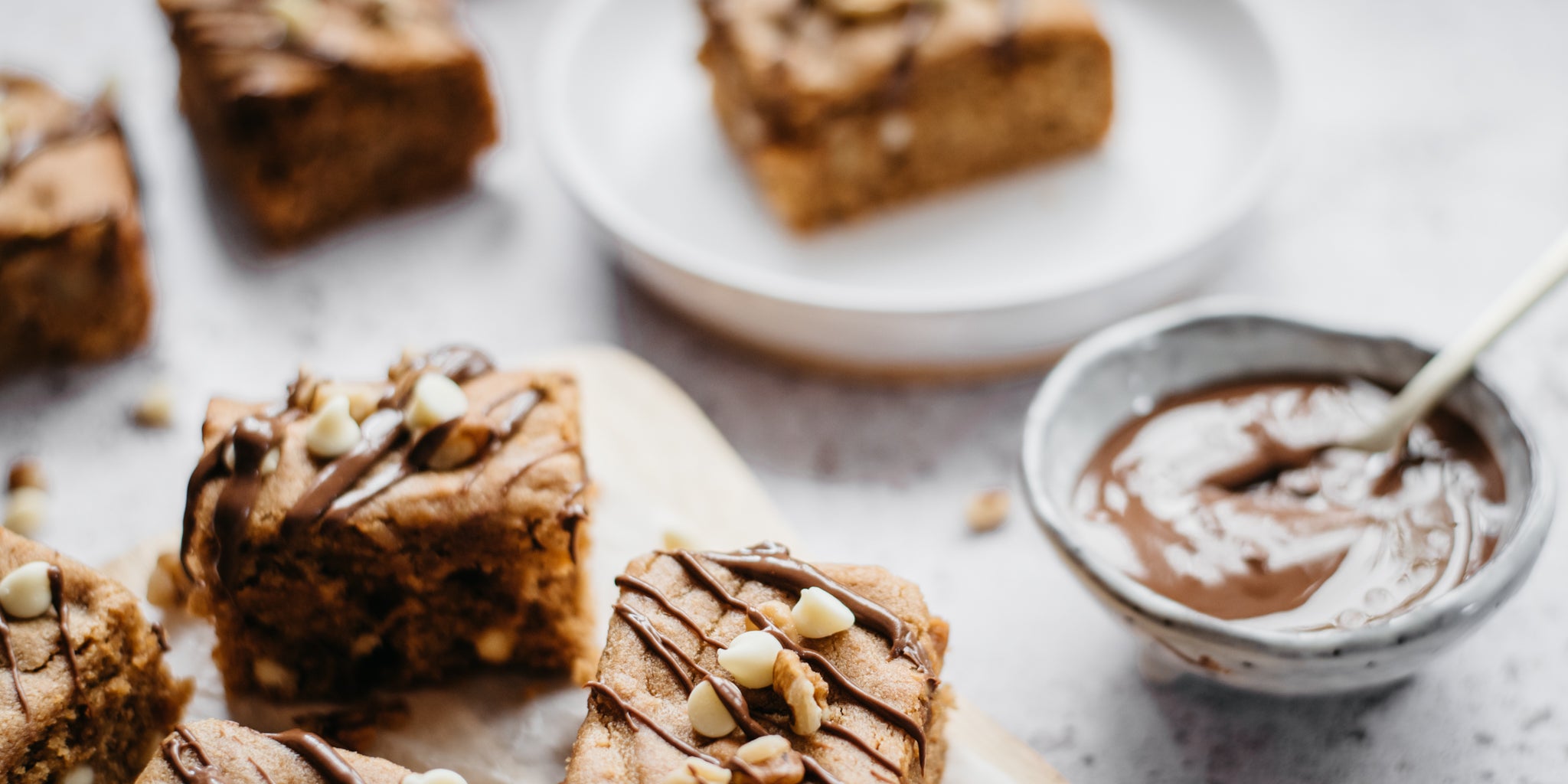 Close up of a peanut butter blondie with white chocolate chips on top and chocolate drizzle. Pot of melted chocolate and spoon in background