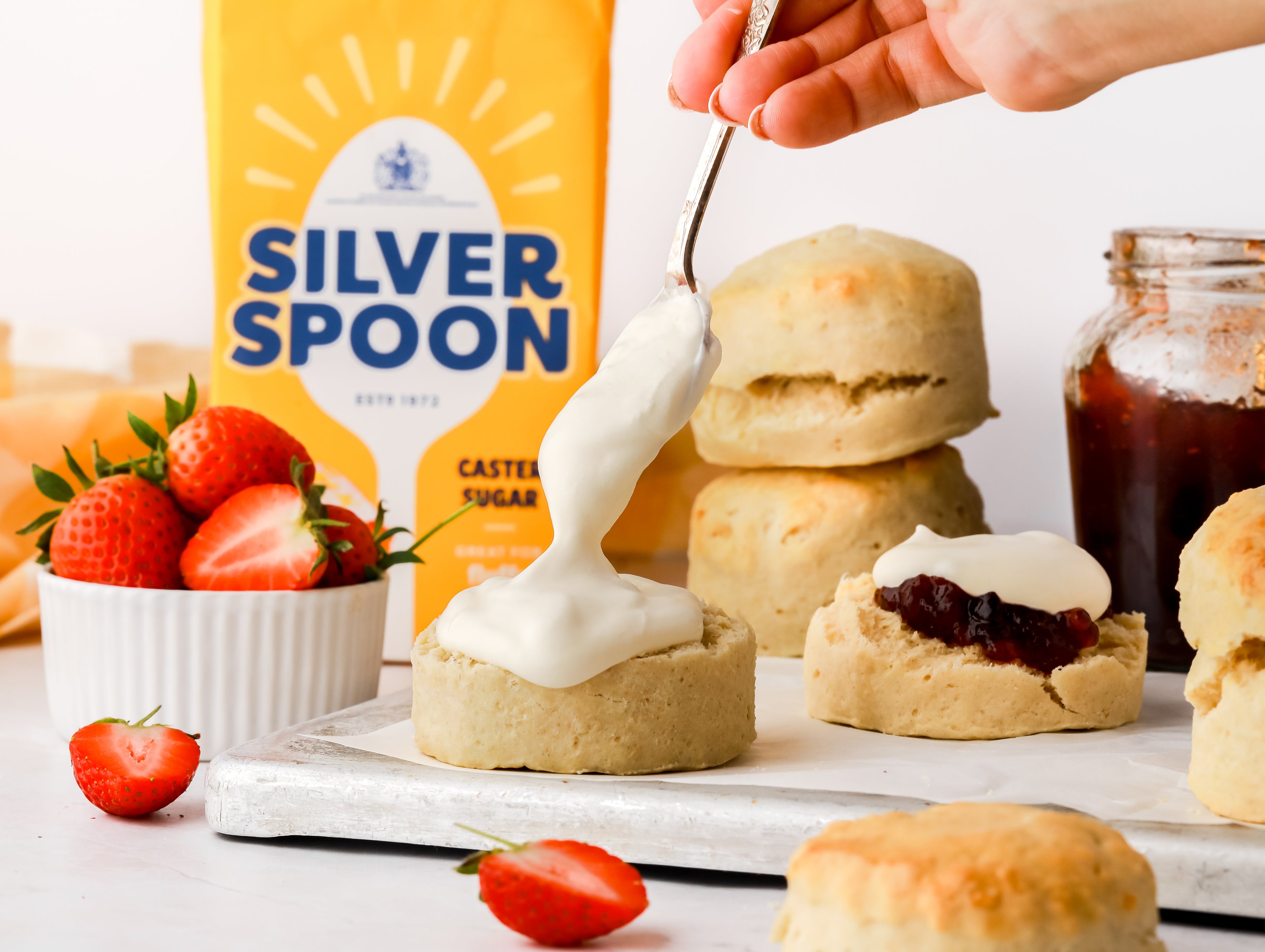 Homemade scone split in two with a dollop of jam and cream from a spoon with strawberries and caster sugar