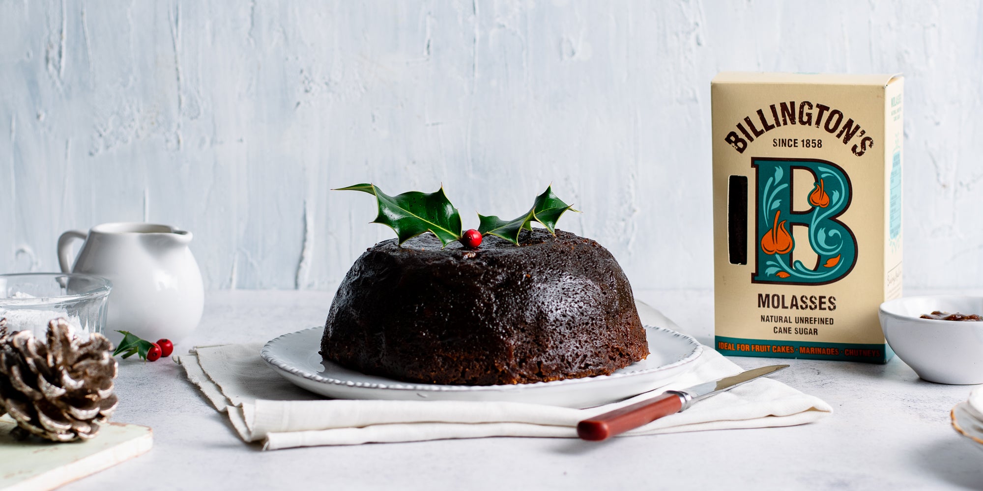 Last Minute Christmas Pudding next to a box of Billington's Molasses and a jug of cream or brandy butter to serve
