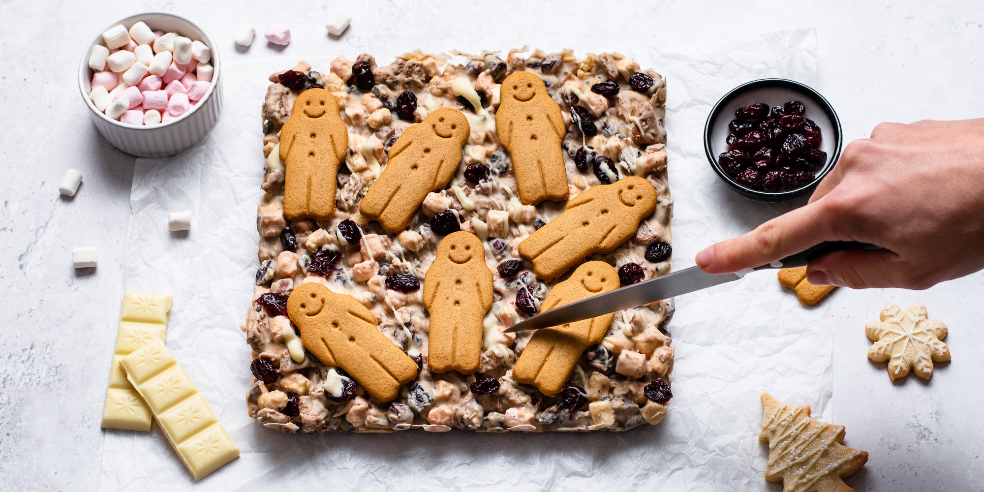 Traybake White Chocolate & Cranberry Rocky Road being slices with a hand holding a knife, next to various toppings of cranberries, marshmallows and white chocolate chunks