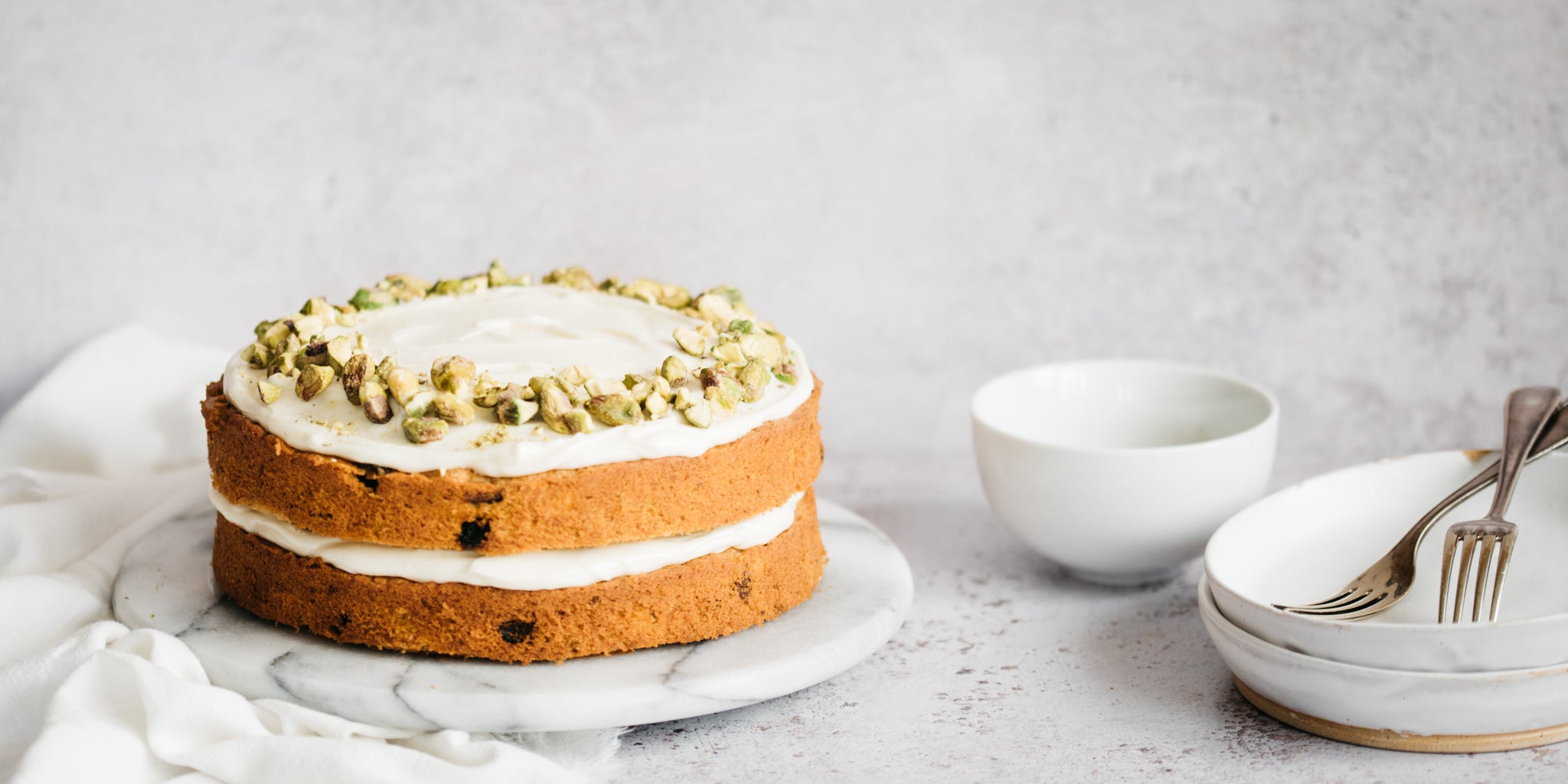 Carrot cake on top of a marble plate next to a stack of white plates, forks and a white bowl