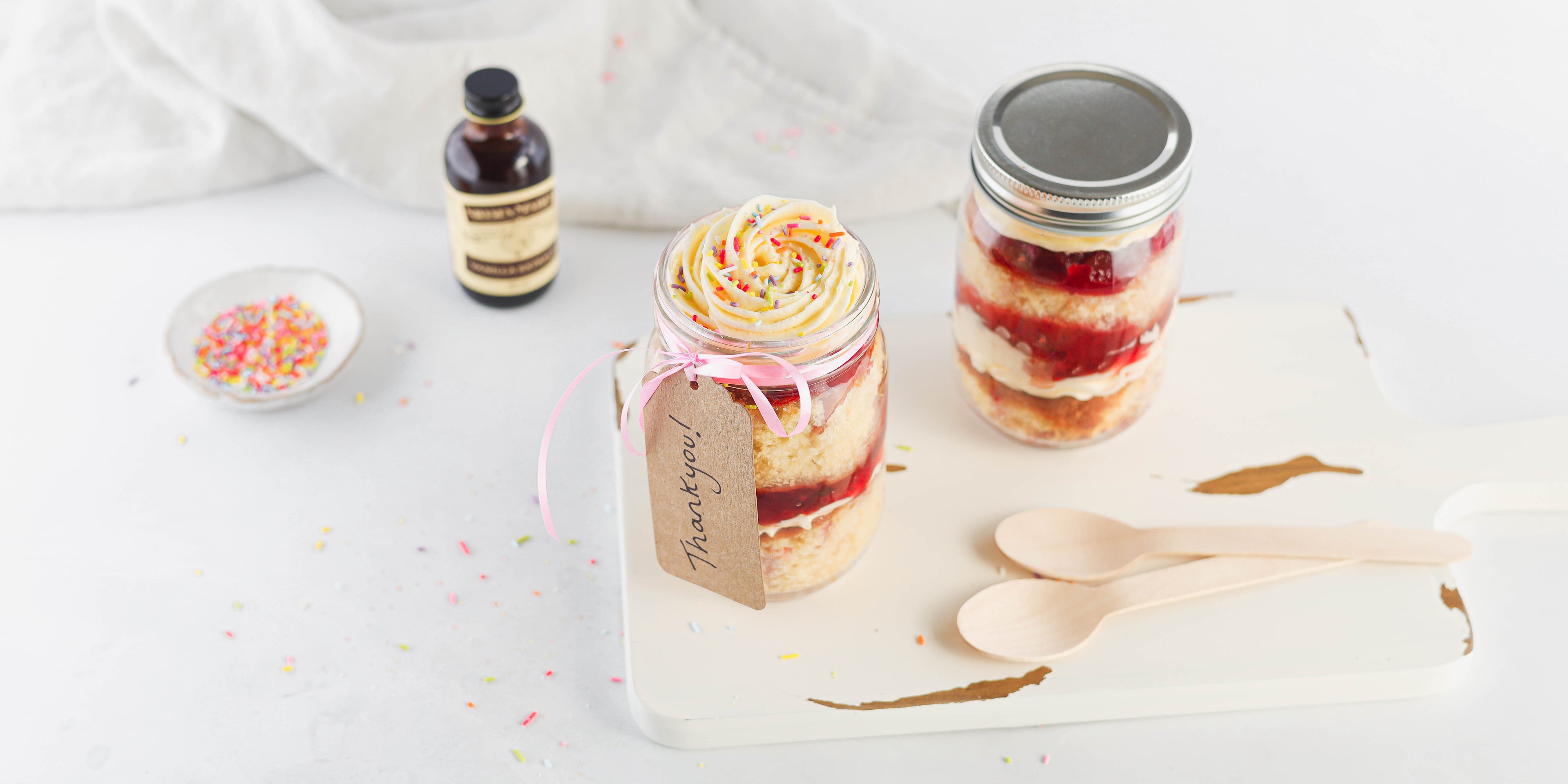 Vanilla Cupcakes in a Jar flat lay. With a bowl of sprinkles, Neilsen-Massey vanilla extract and a handwritten thank you label