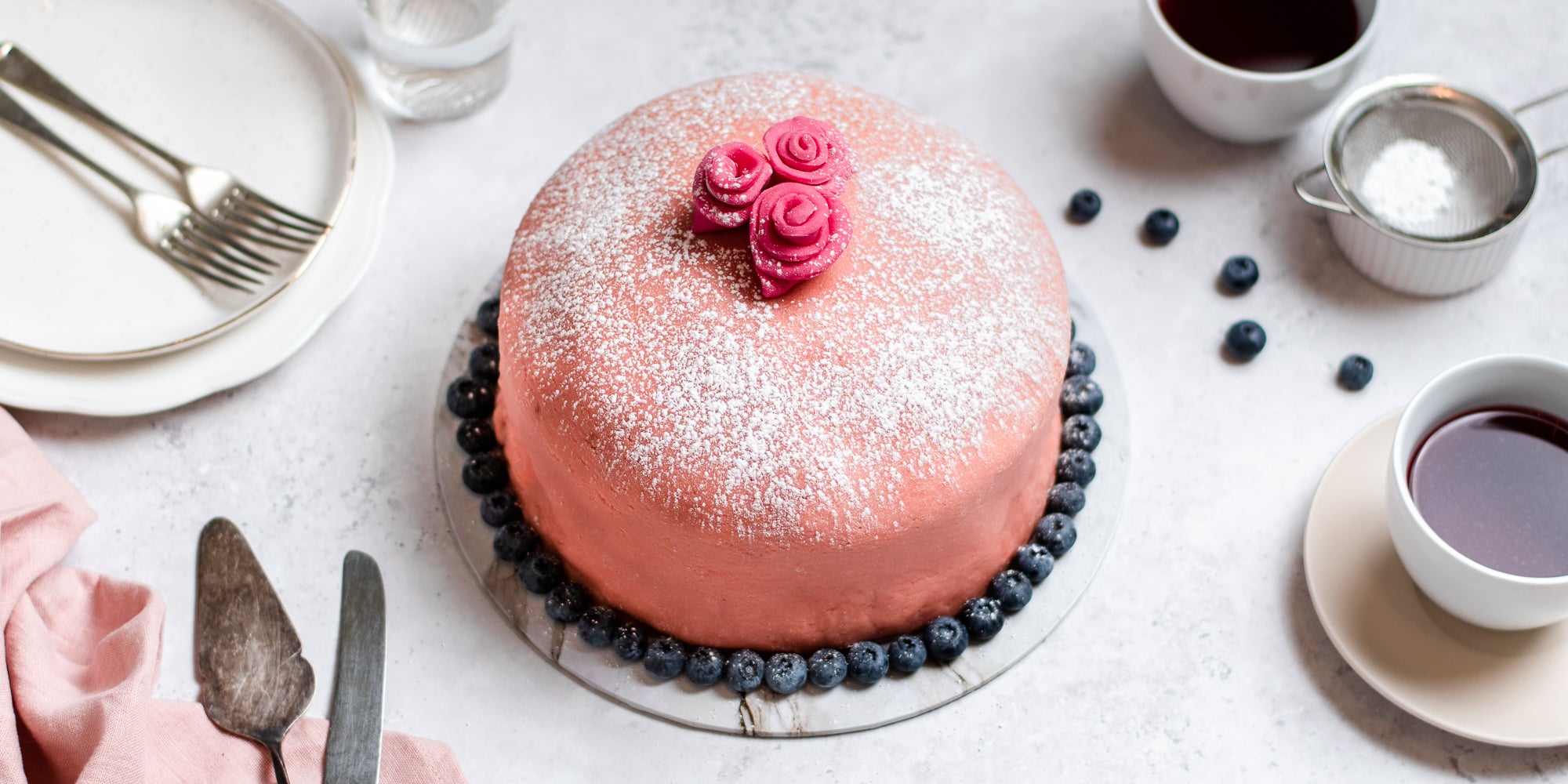 Prinsesstarta surrounded by blueberries, next to cups and saucers and a sieve. There is a cake knife to serve, with plates and cutlery