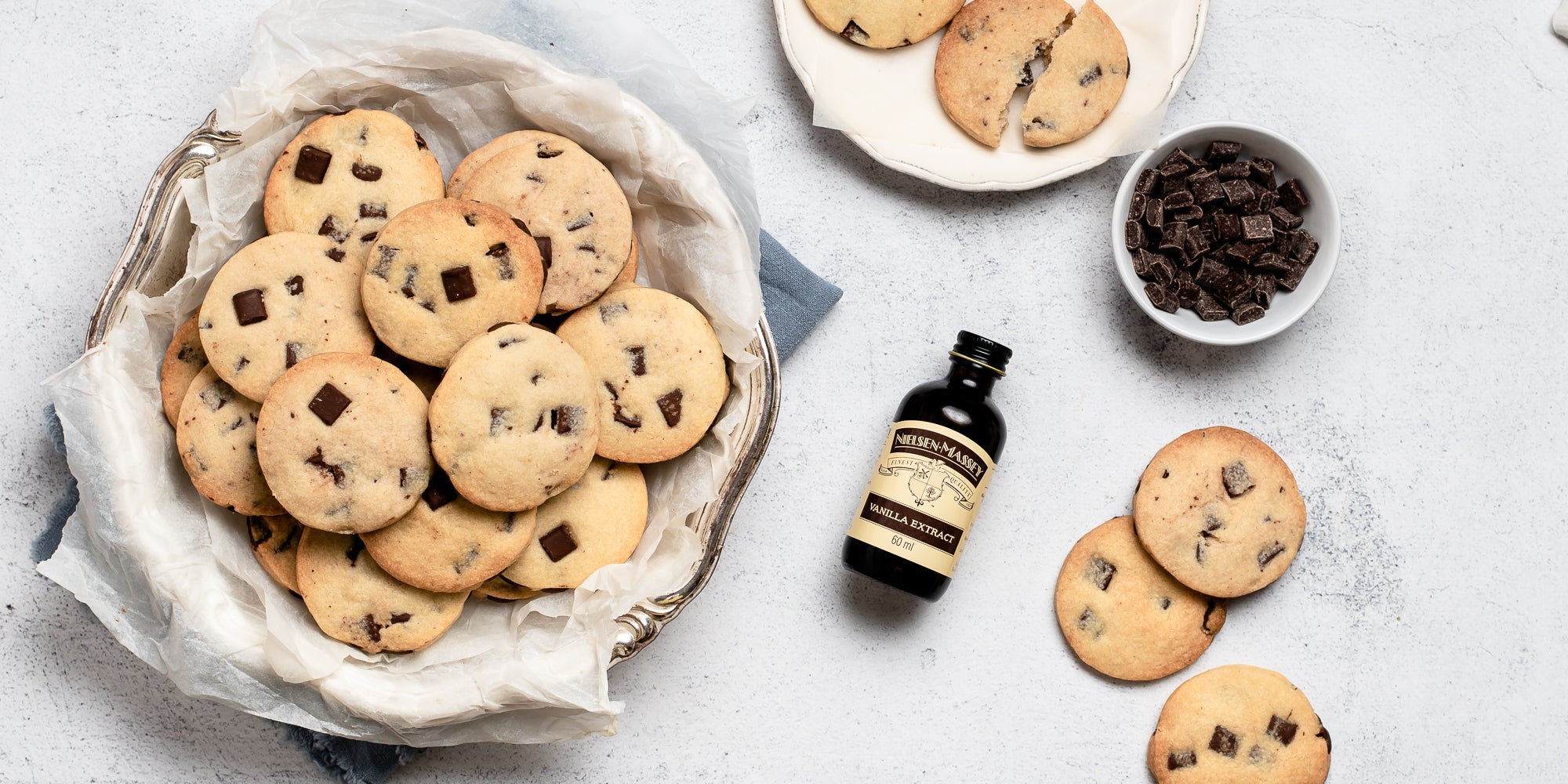 Top view of Chocolate Chunk Shortbread in a cookie tin, next to a bottle of Nielsen-Massey vanilla extract and some shortbread biscuits