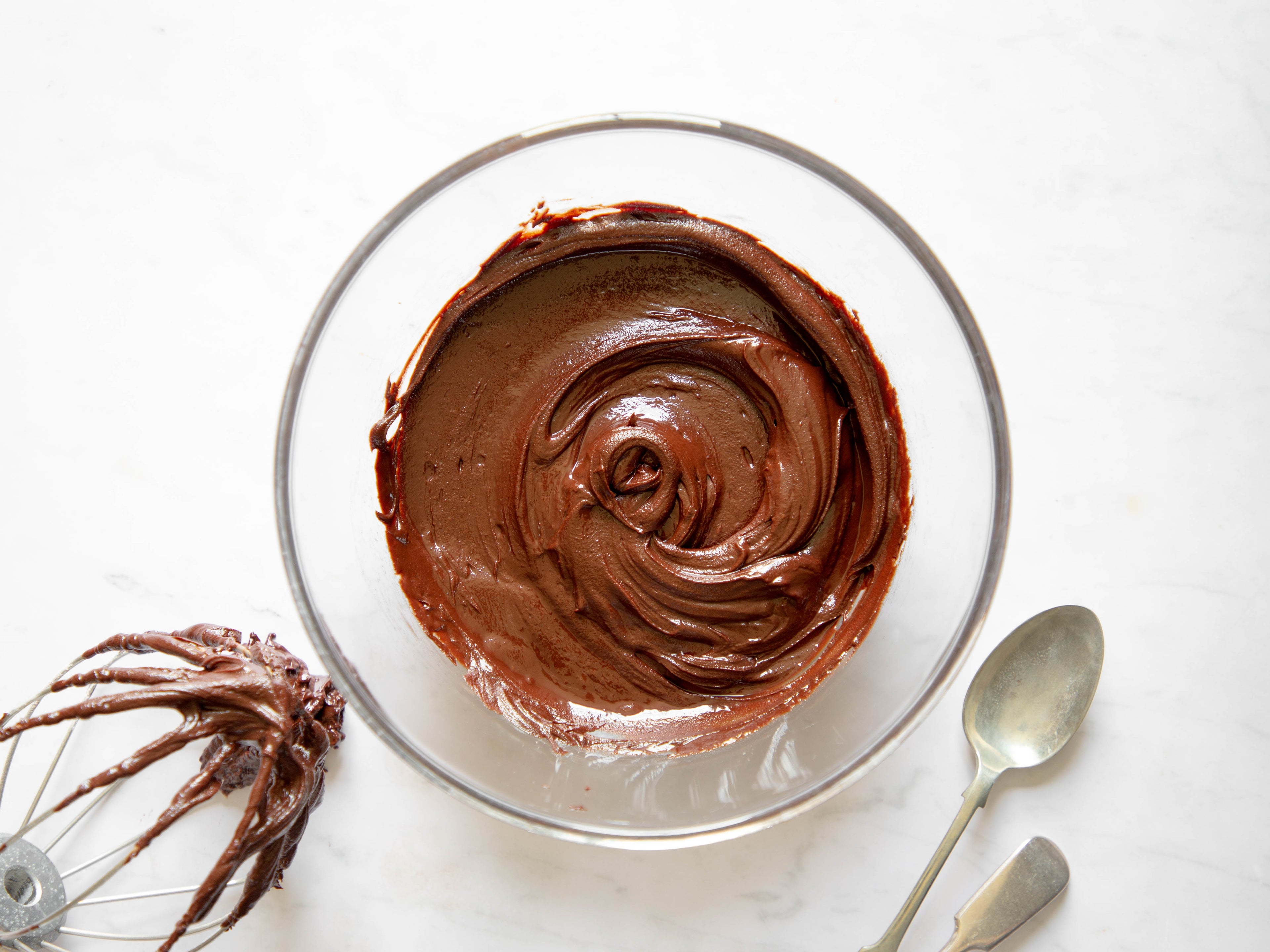 Glass Bowl of chocolate icing with a chocolate covered whisk on the side to the left and two spoons on the right