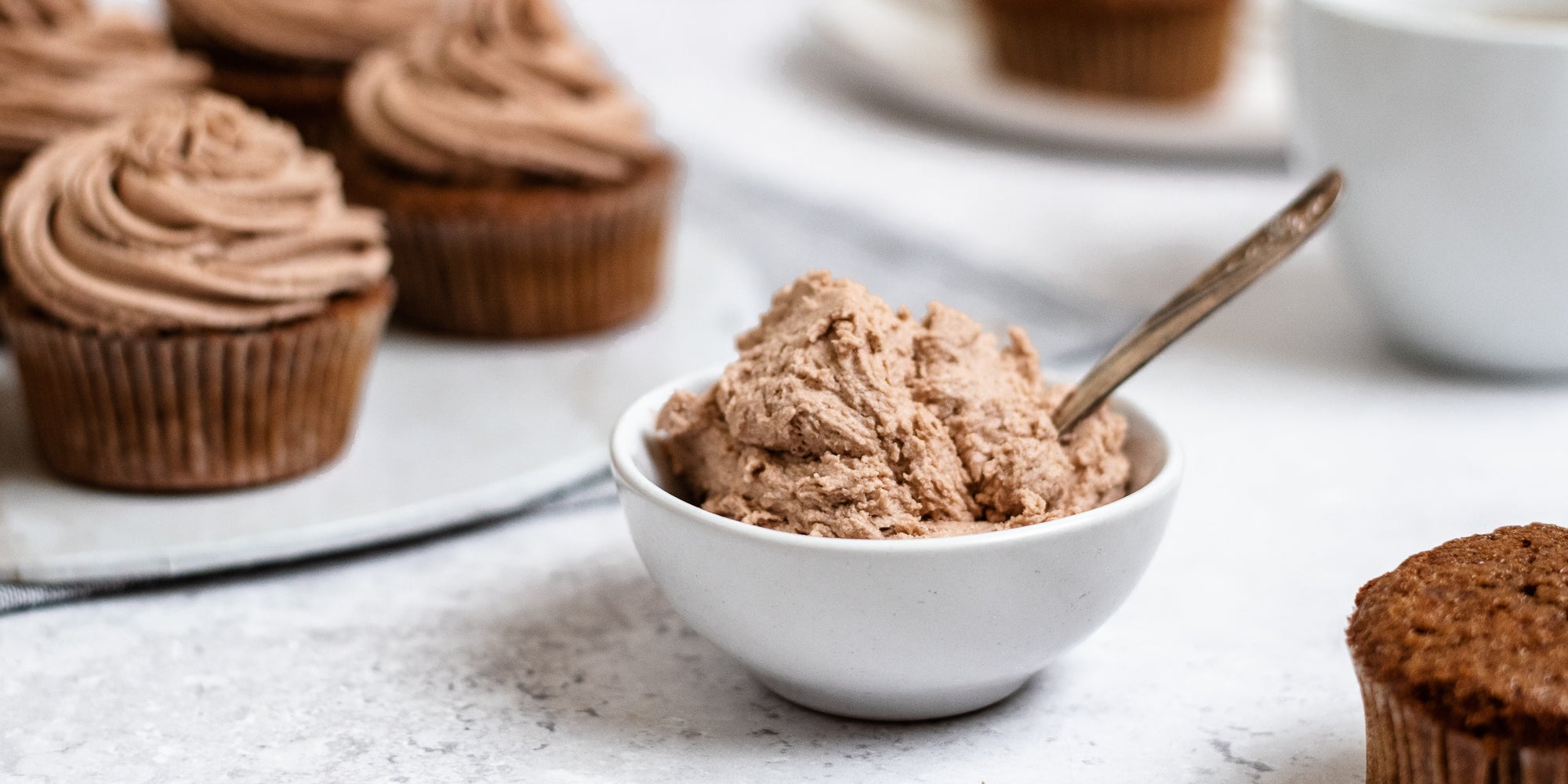 Close up of Dairy Free Vegan Chocolate Buttercream in a small bowl, with a spoon. Cupcakes topped with Dairy Free Vegan Chocolate Buttercream in the background