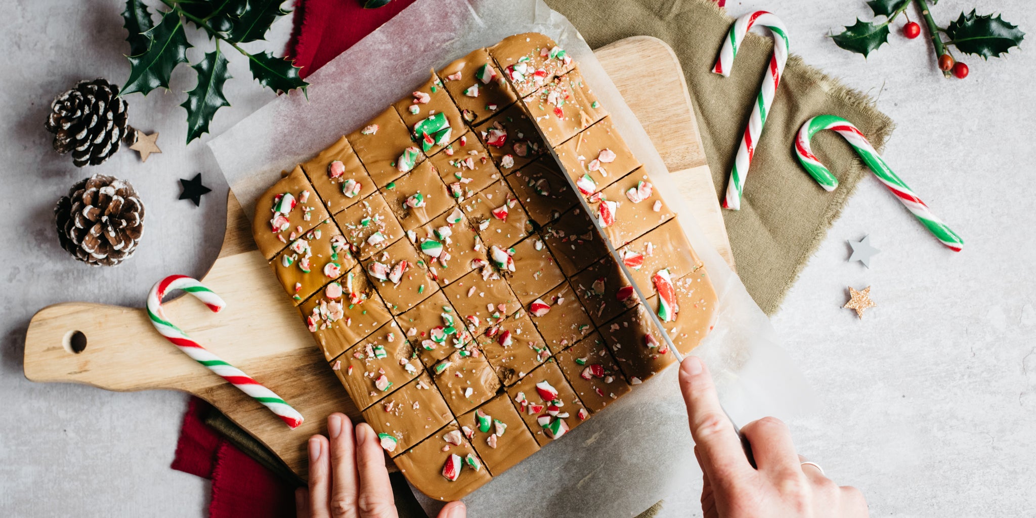 Top view of Billington's Candy Cane Fudge being chopped into squares by a hand. On a wooden chopping board next to candy canes and pine cones.