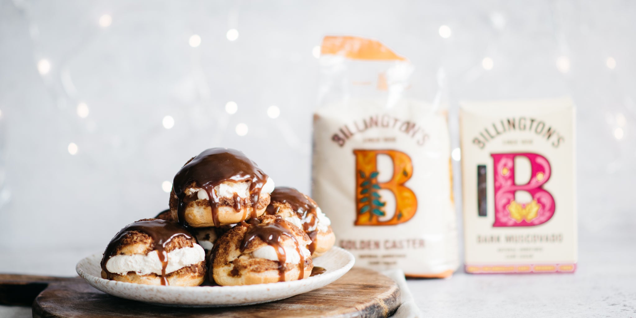 A plate of profiteroles stacked with a toffee sauce, two sugar packs in background with fairy lights