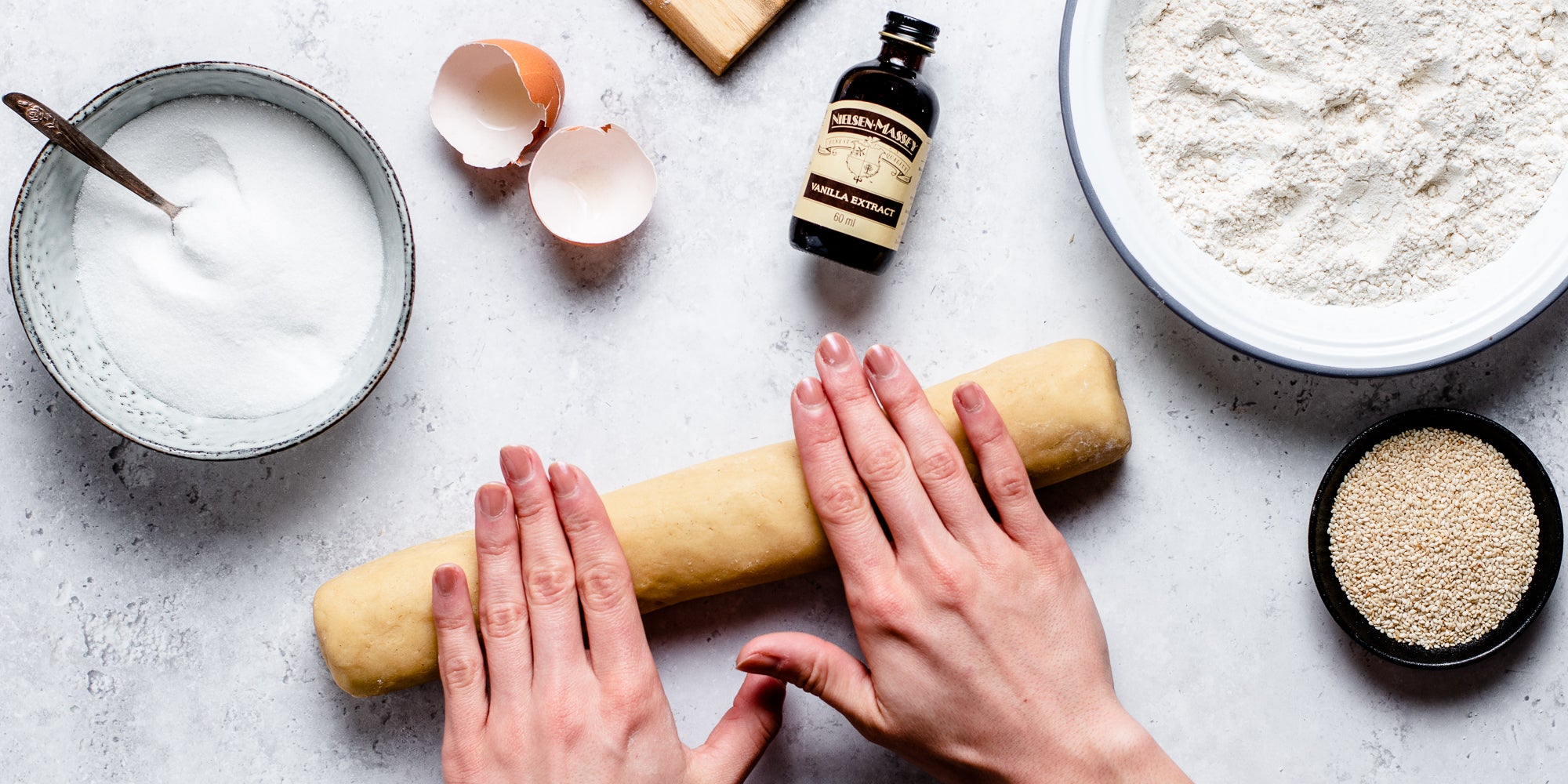 Top down view of some hands rolling a rolling pin surrounded by sesame cookie ingredients
