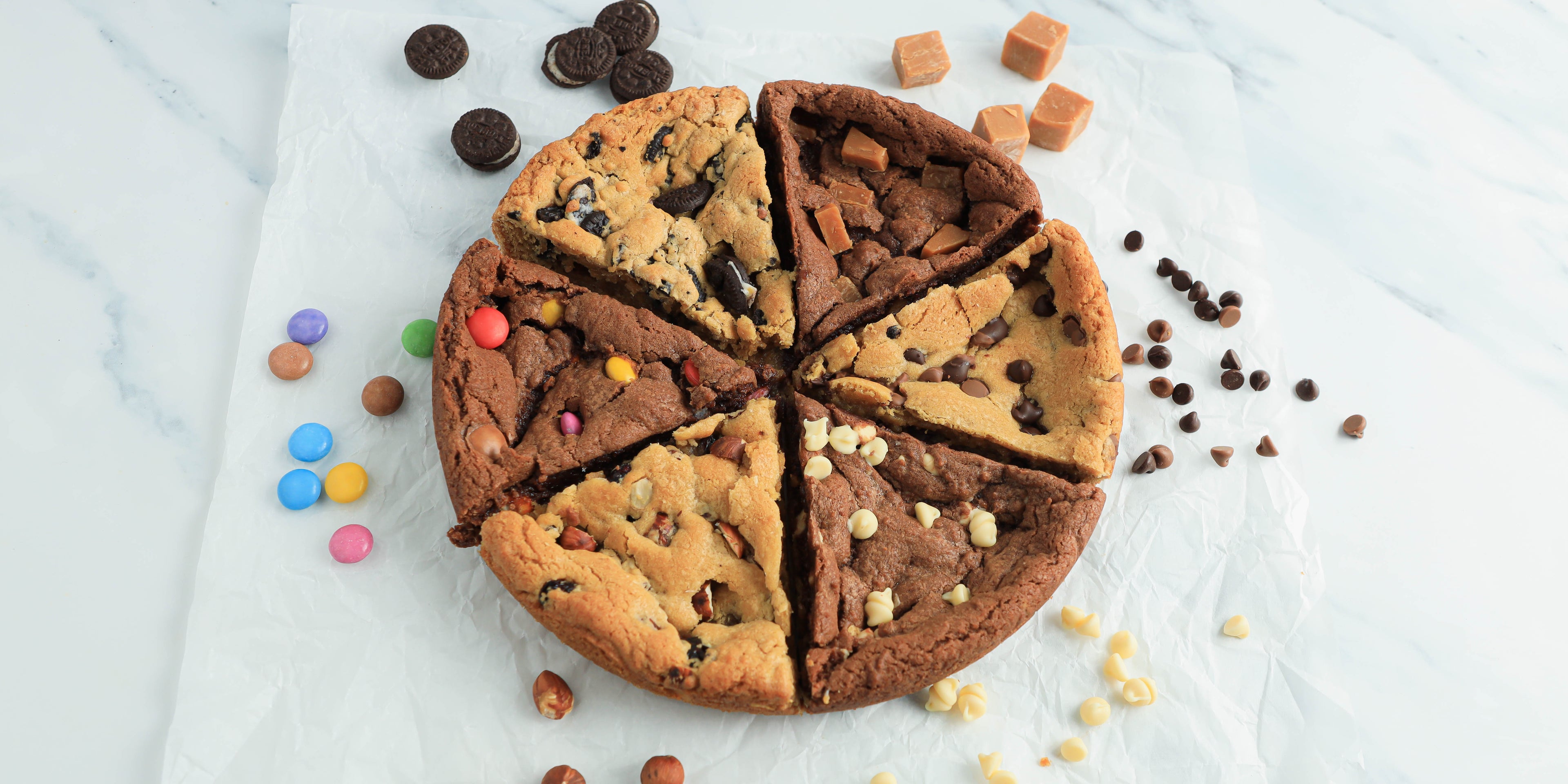 Cookie cake cut into 6 slices with sweets, biscuits and chocolate chips scattered around the outside