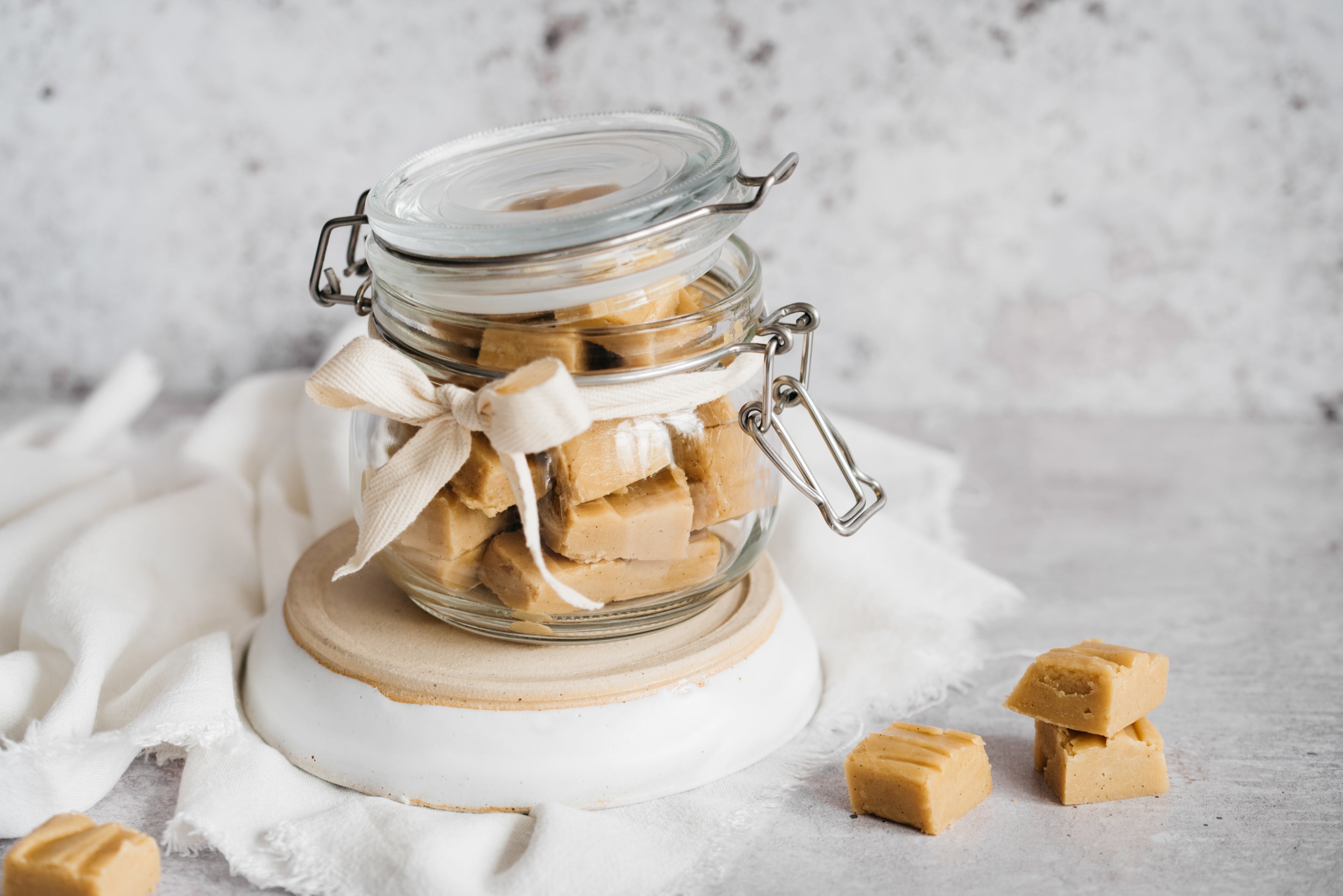 Cubes of vanilla fudge in an open kilner jar decorated with a cream ribbon bow. 4 cubes of fudge in the forefront