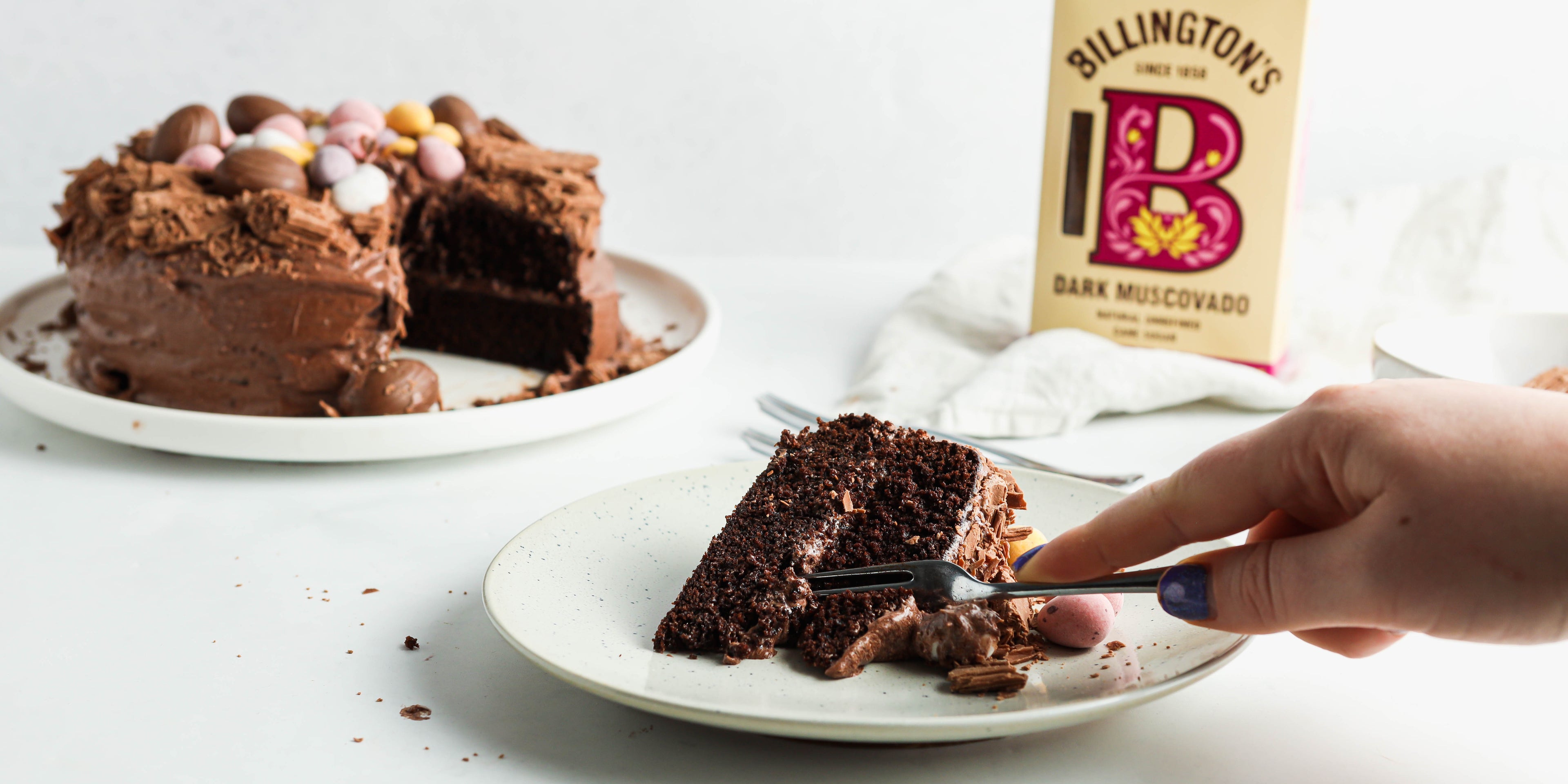Chocolate Nest Cake slice being cut into with a hand holding a fork, with a box of Billington's Dark Muscovado sugar in the background