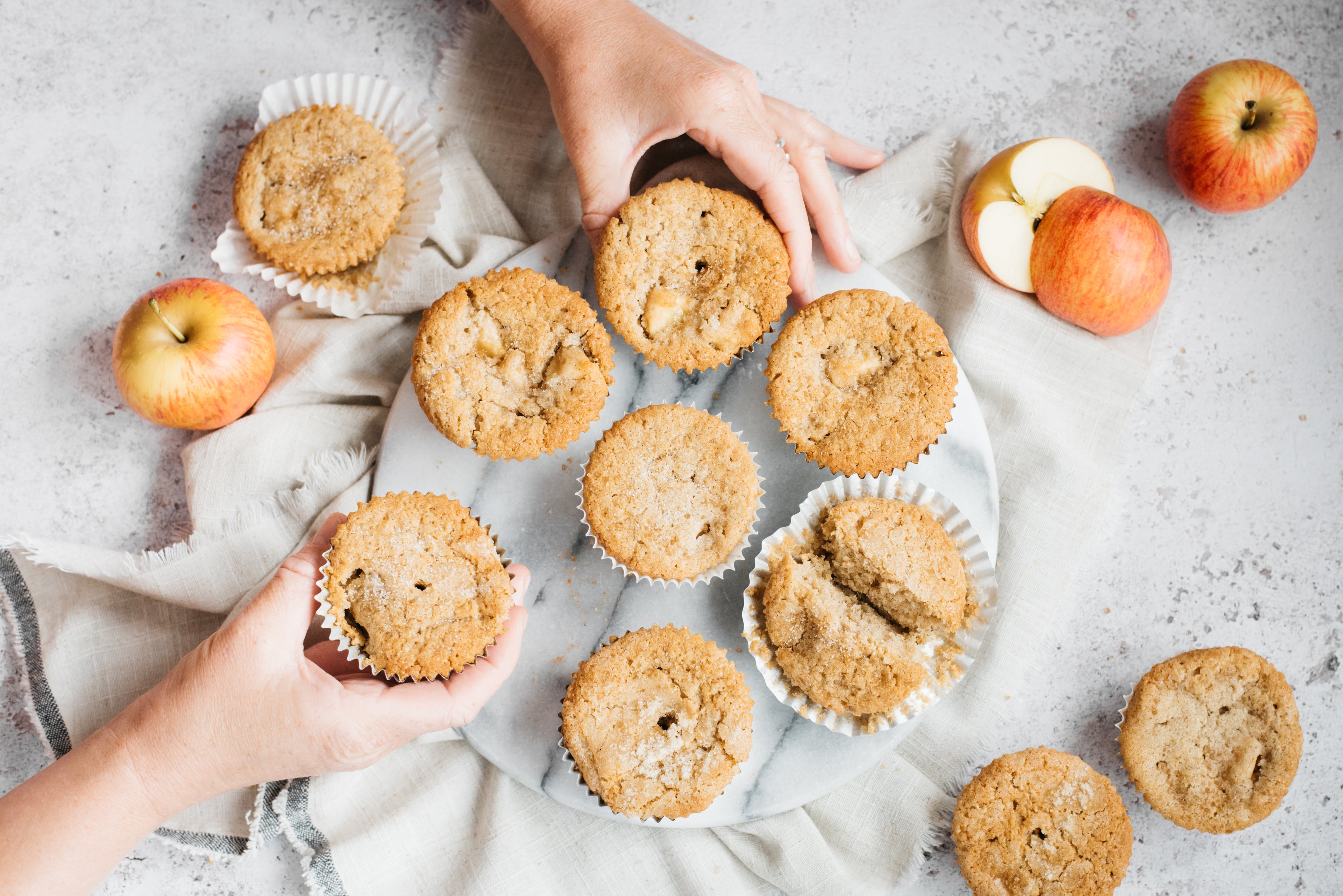Hands reaching in to grab a plate of muffins with apples beside