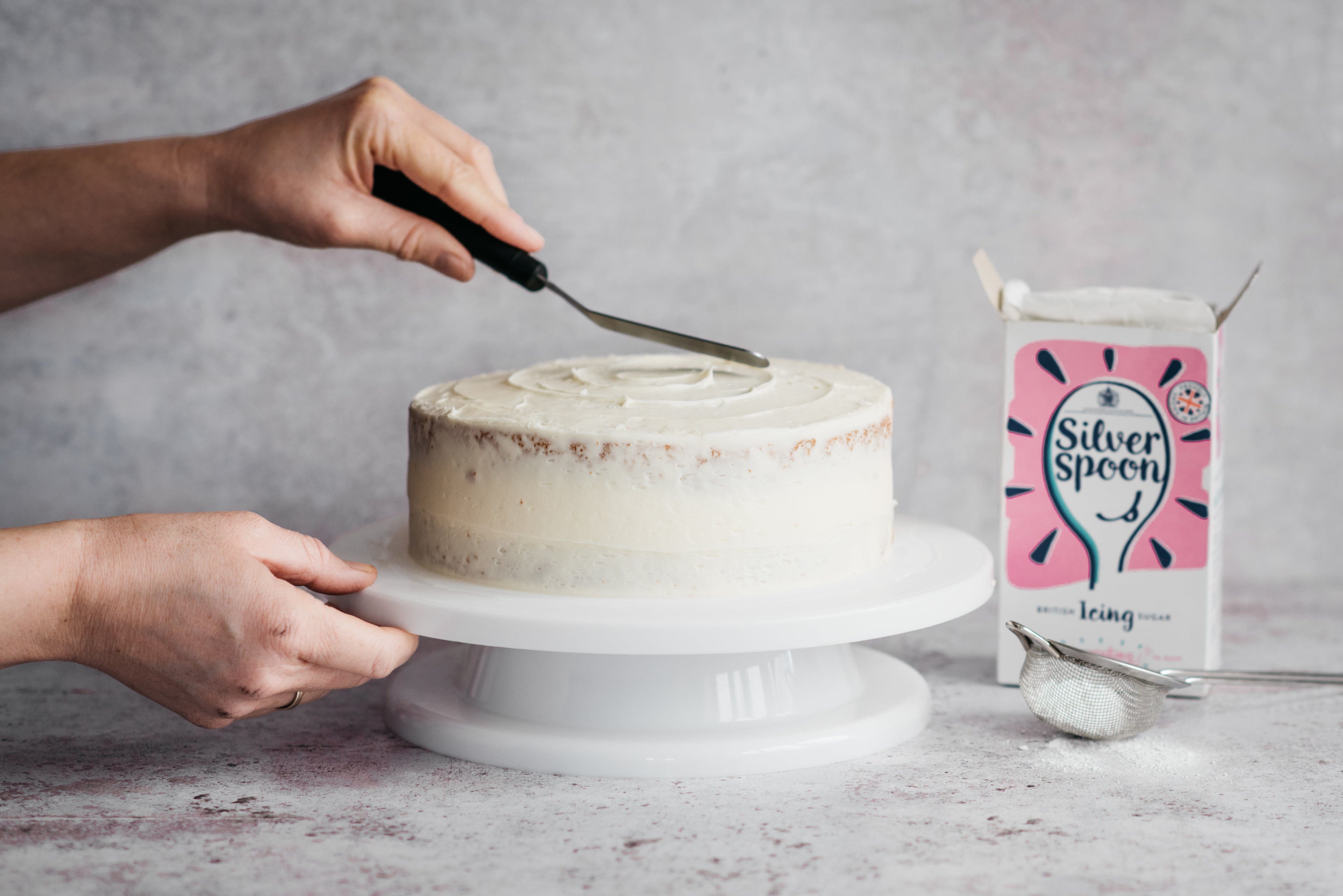 Hand using an offset spatula to smooth a layer of Silver Spoon Buttercream over the Gluten Free Elderflower & Lemon Cake