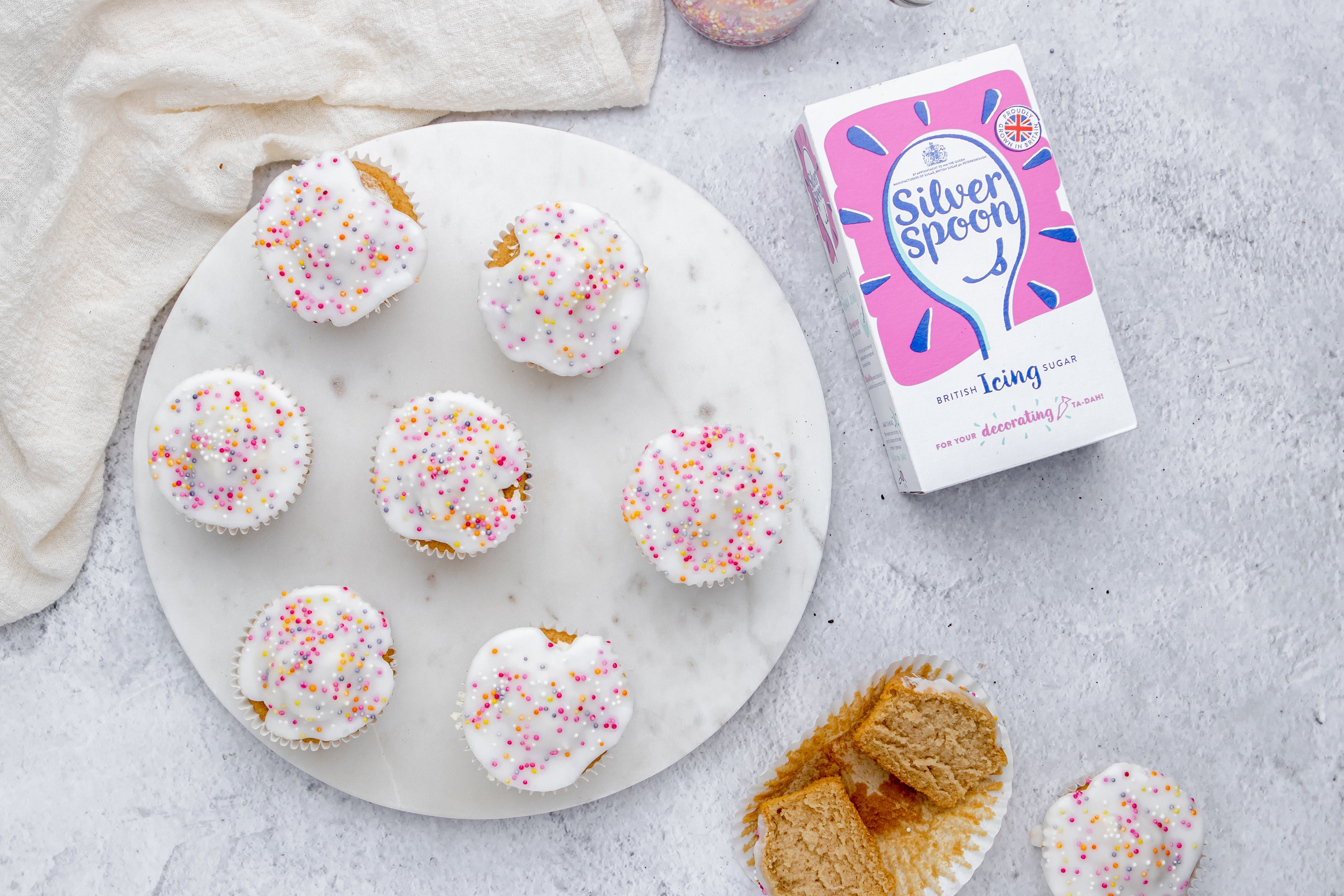 Simple Fairy Cakes on a plate next to a box of Silver Spoon Icing Sugar, and a sliced cupcake in a cupcake case