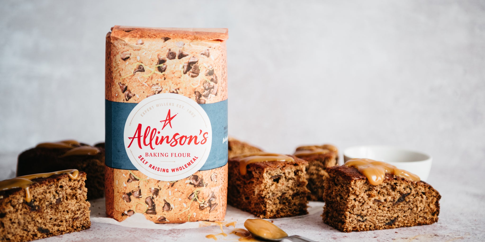 Ultimate Wholemeal Sticky Toffee Pudding slices drizzled with toffee sauce on parchment paper, next to a bag of Allinson's Wholemeal Flour