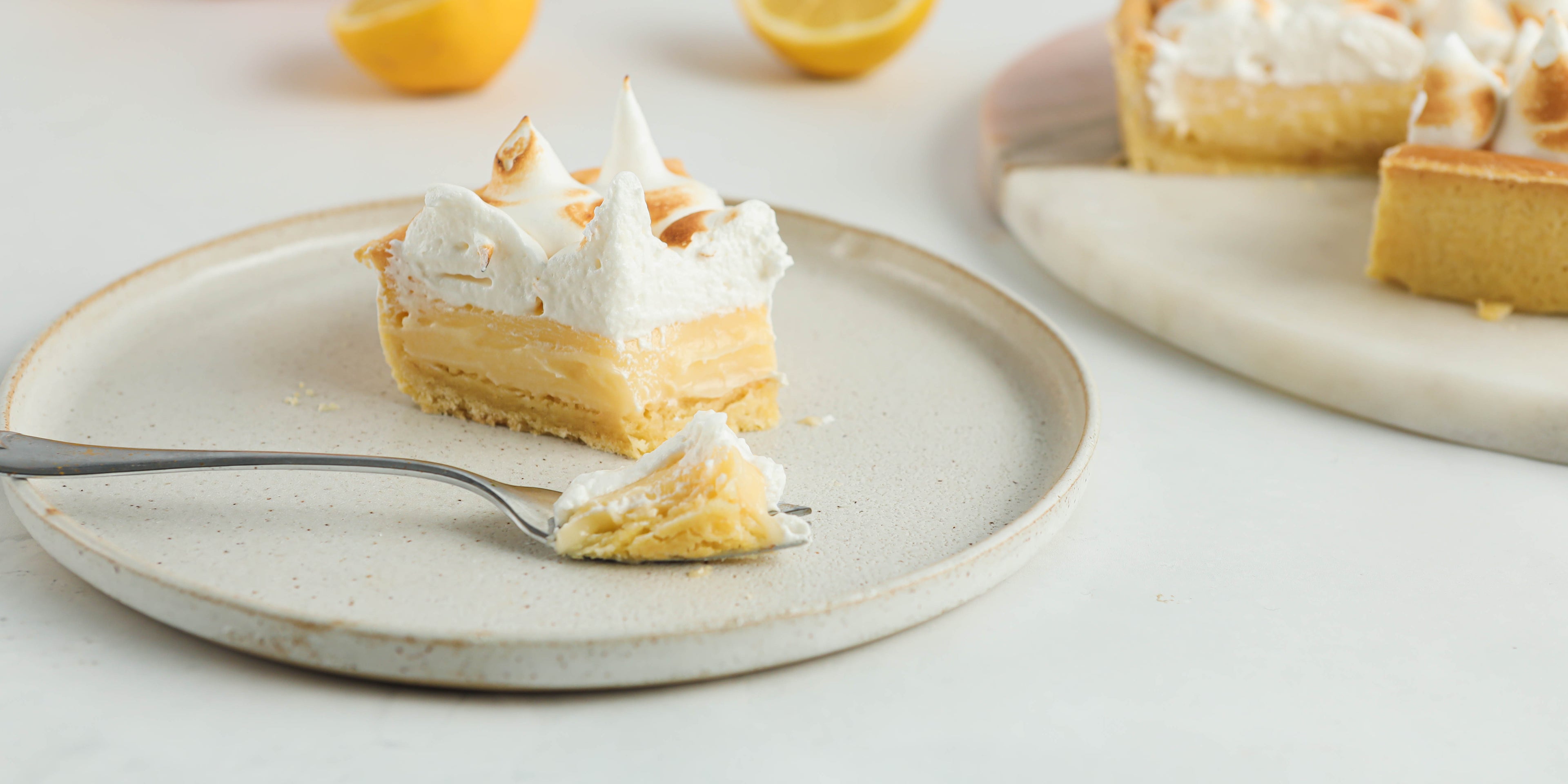 A slice of lemon meringue pie on a white plate with a forkful of pie infront. The remaining pie is in the background on a white plate with some spare lemons beside it.