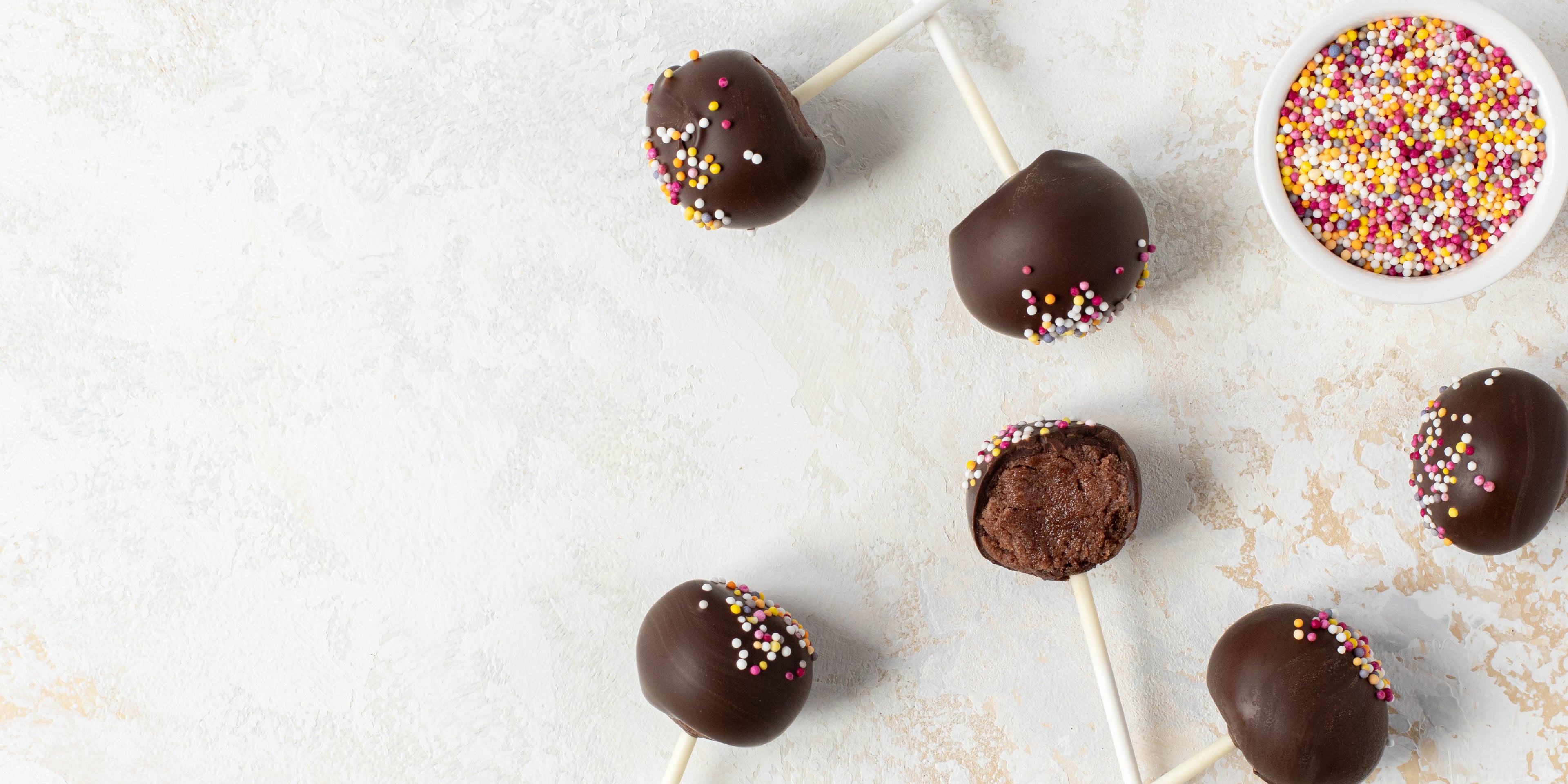 Top view of Chocolate Cake Pops scattered with a bite taken out of a cake pop, showing the centre, next to a bowl of sprinkles.