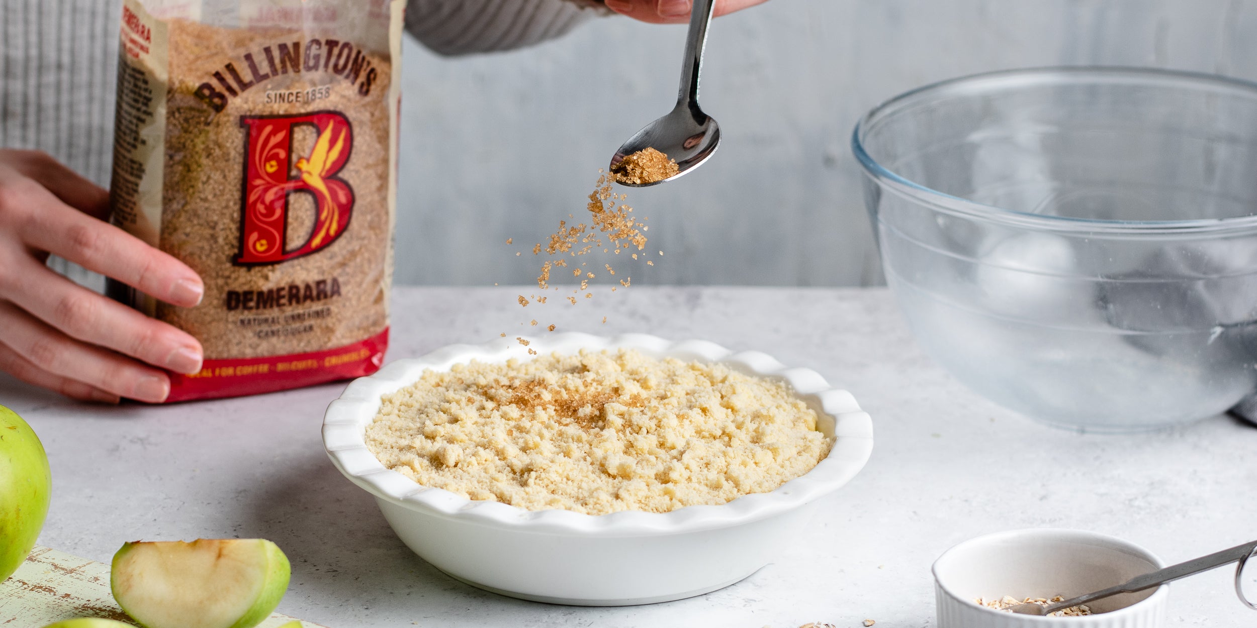 Chopped apples being sprinkled with crumble mixture ready to bake. Next to a bag of Billington's Demerara sugar