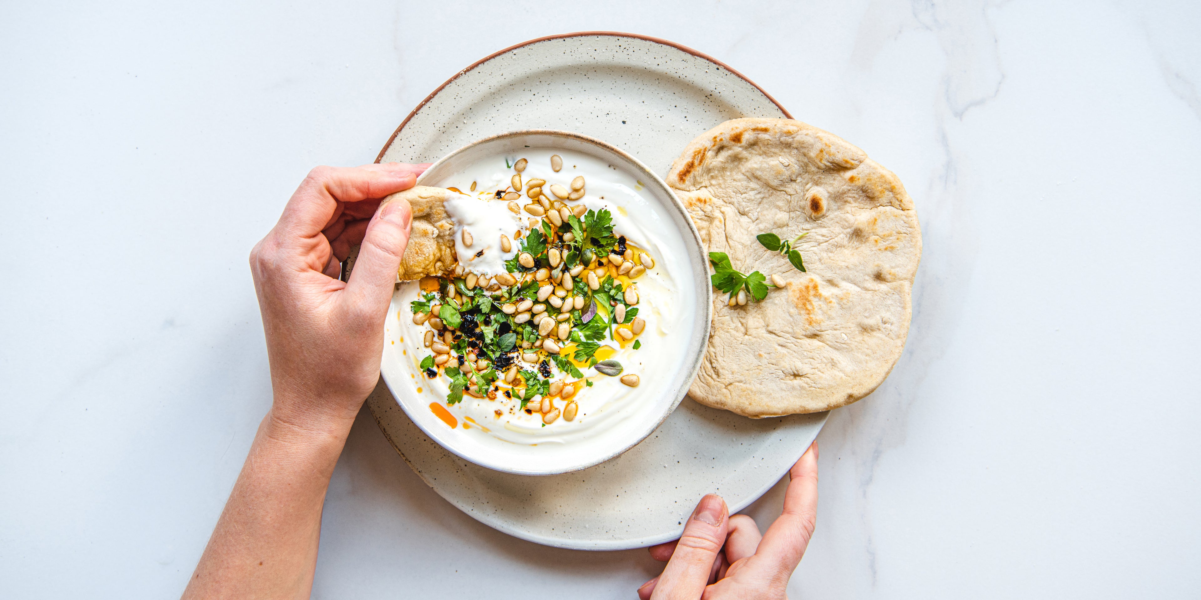 A top down view of a plate with a hand dipping a piece of Kefir flatbread into a bowl of tzatziki