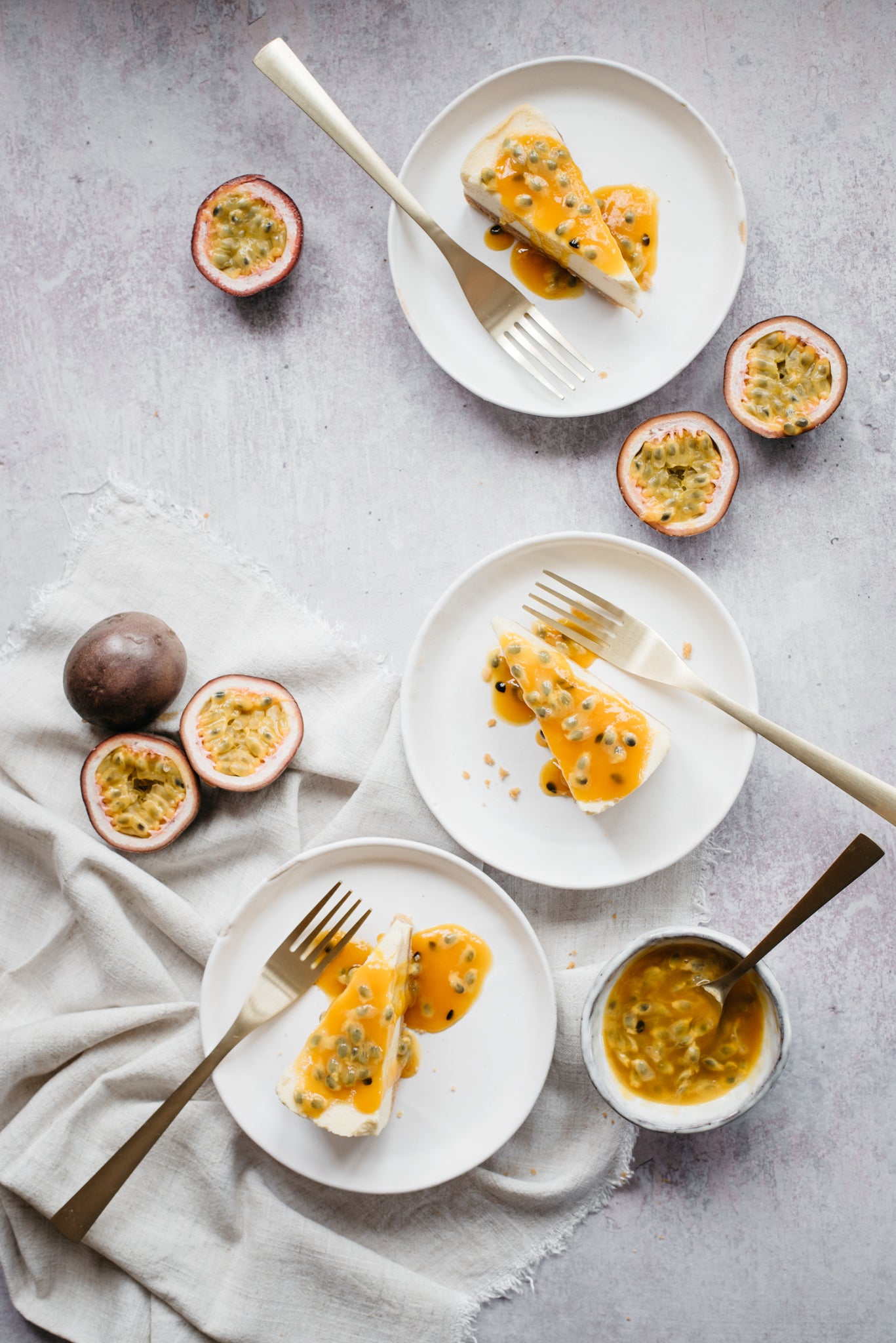 Overhead shot of 3 plates each with a slice of passionfruit cheesecake and a fork. 3 passionfruits beside the plate and a bowl of passion fruit sauce
