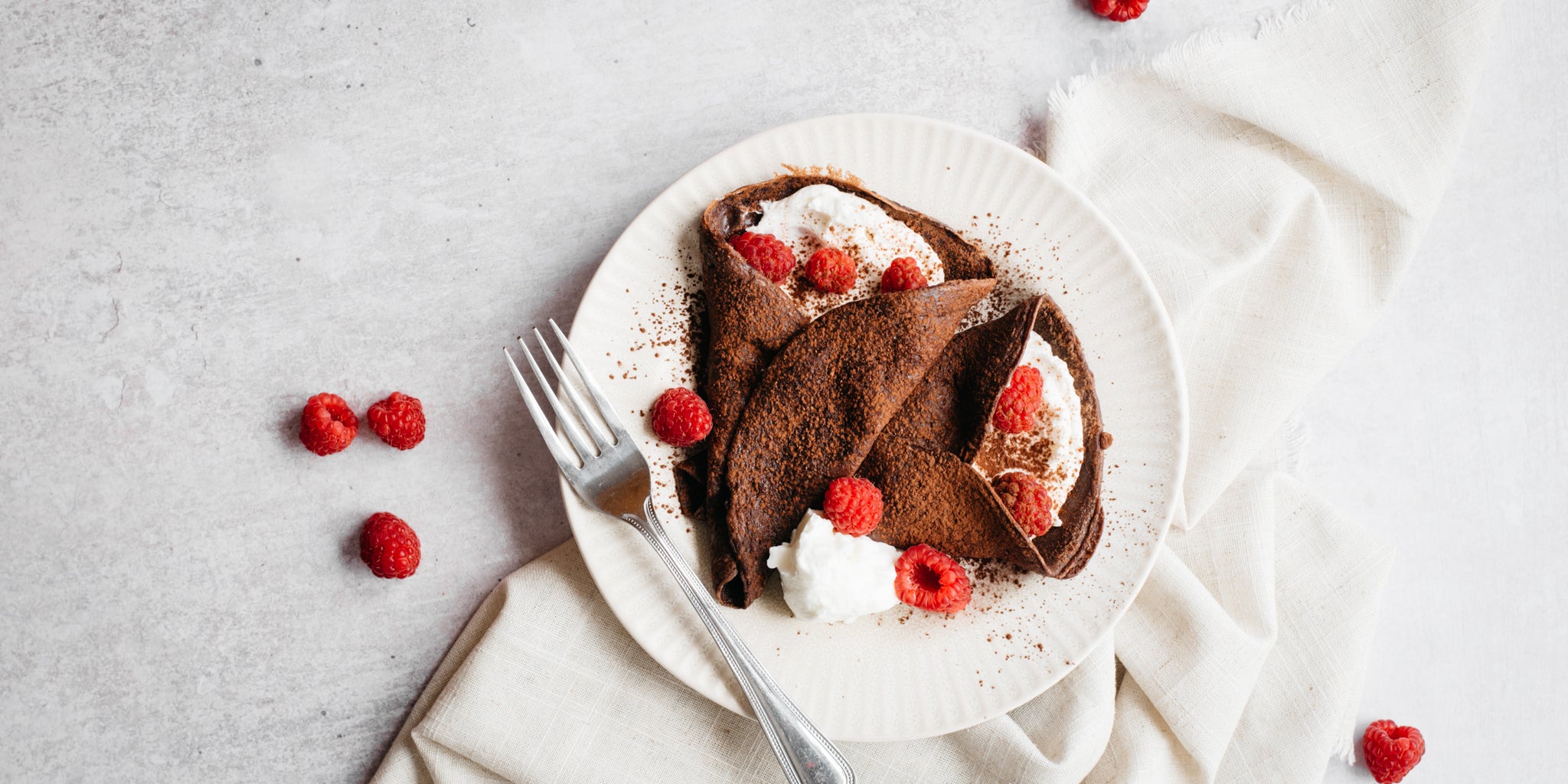 White plate with two chocolate crepes with cream and raspberries