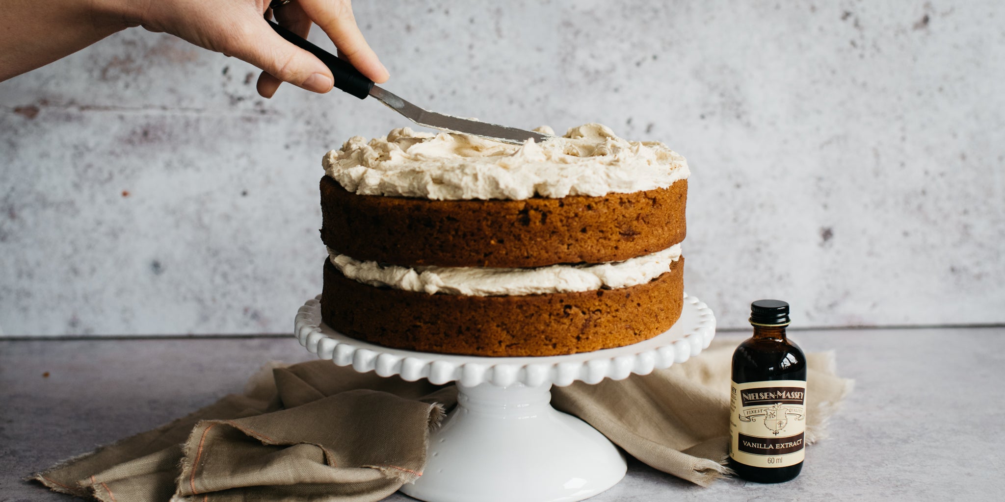 Vegan Carrot Cake being sliced into using a cake knife, next to a bottle of Nielsen-Massey vanilla extract